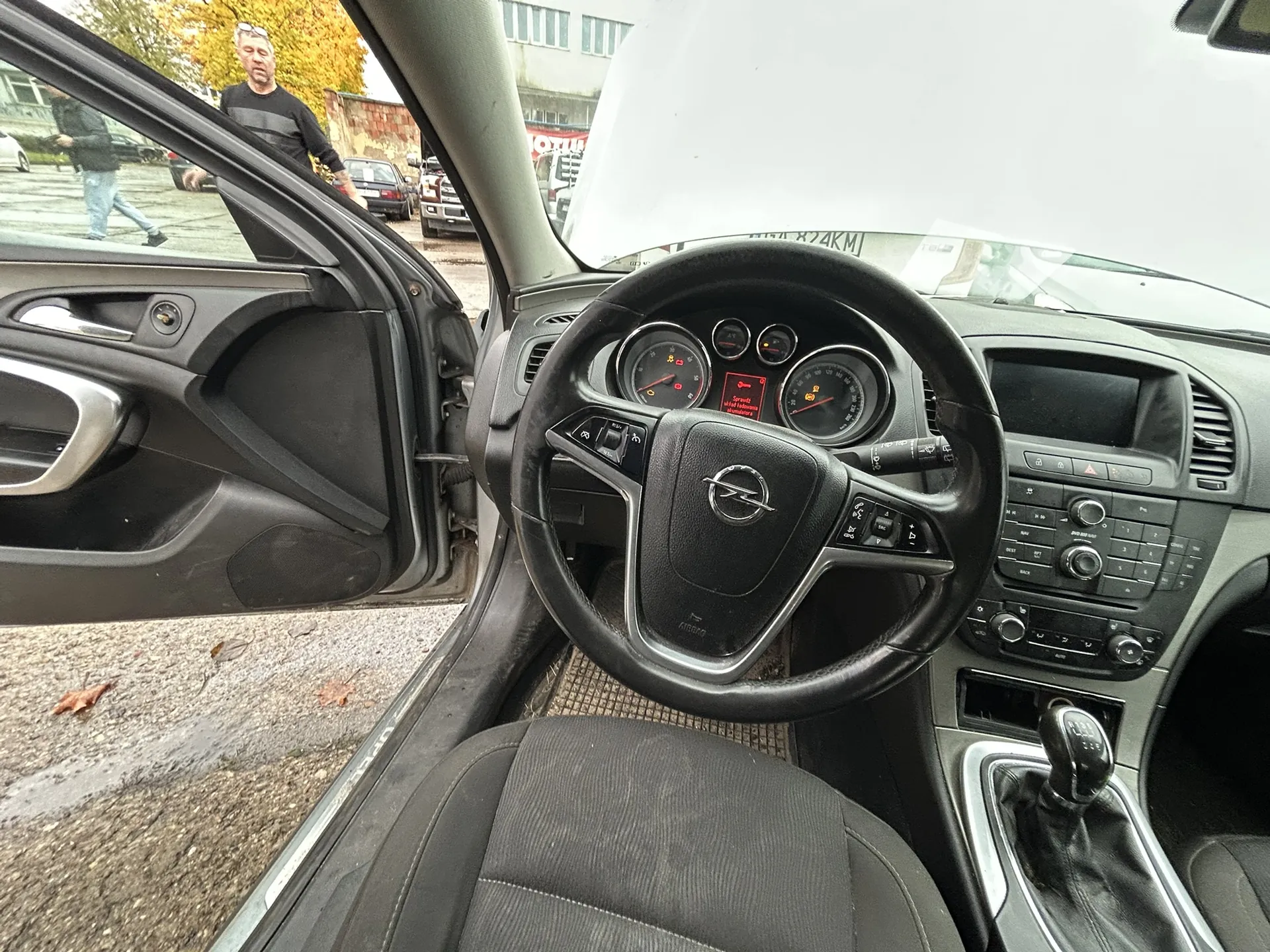 Wnętrze samochodu Opel z widocznym kierownicą, tablicą przyrządów i centralną konsolą. Drzwi po stronie kierowcy są otwarte, na zewnątrz widać mężczyznę stojącego na mokrej nawierzchni, inne pojazdy oraz drzewa z jesiennymi liśćmi. Tablica przyrządów wyświetla wskaźniki prędkości i obrotomierza, a na środku kierownicy znajduje się logo Opel. W tle widoczne są budynki i zaparkowane samochody.