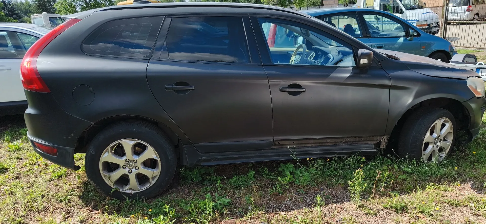 Czarny samochód typu crossover zaparkowany na trawiastym terenie. Pojazd jest widoczny z profilu, z boku. W tle widoczne są inne samochody oraz fragment ogrodzenia.
