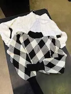 Czarno-biały sweter w argyle z białym ubraniem pod spodem leżący na ciemnej powierzchni. Tło z brązową podłogą i częścią czarnego przedmiotu.