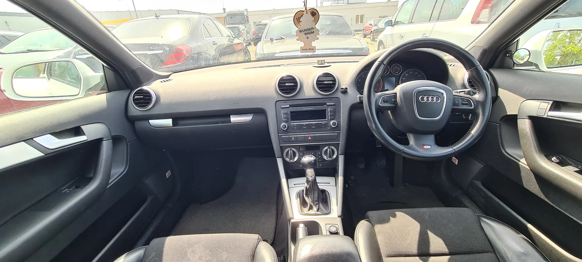 Wnętrze samochodu marki Audi z widocznym kierownicą z logo Audi, centralną konsolą z przyciskami i ekranem, dźwignią zmiany biegów oraz czarnymi siedzeniami. Na desce rozdzielczej wisząca ozdoba z napisem "WUNDER-BAUM Arctic White". Przez przednią szybę widoczne są zaparkowane samochody na zewnątrz. Wnętrze posiada szare i czarne elementy, a po bokach widoczne są drzwi z uchwytami i lusterkami bocznymi.