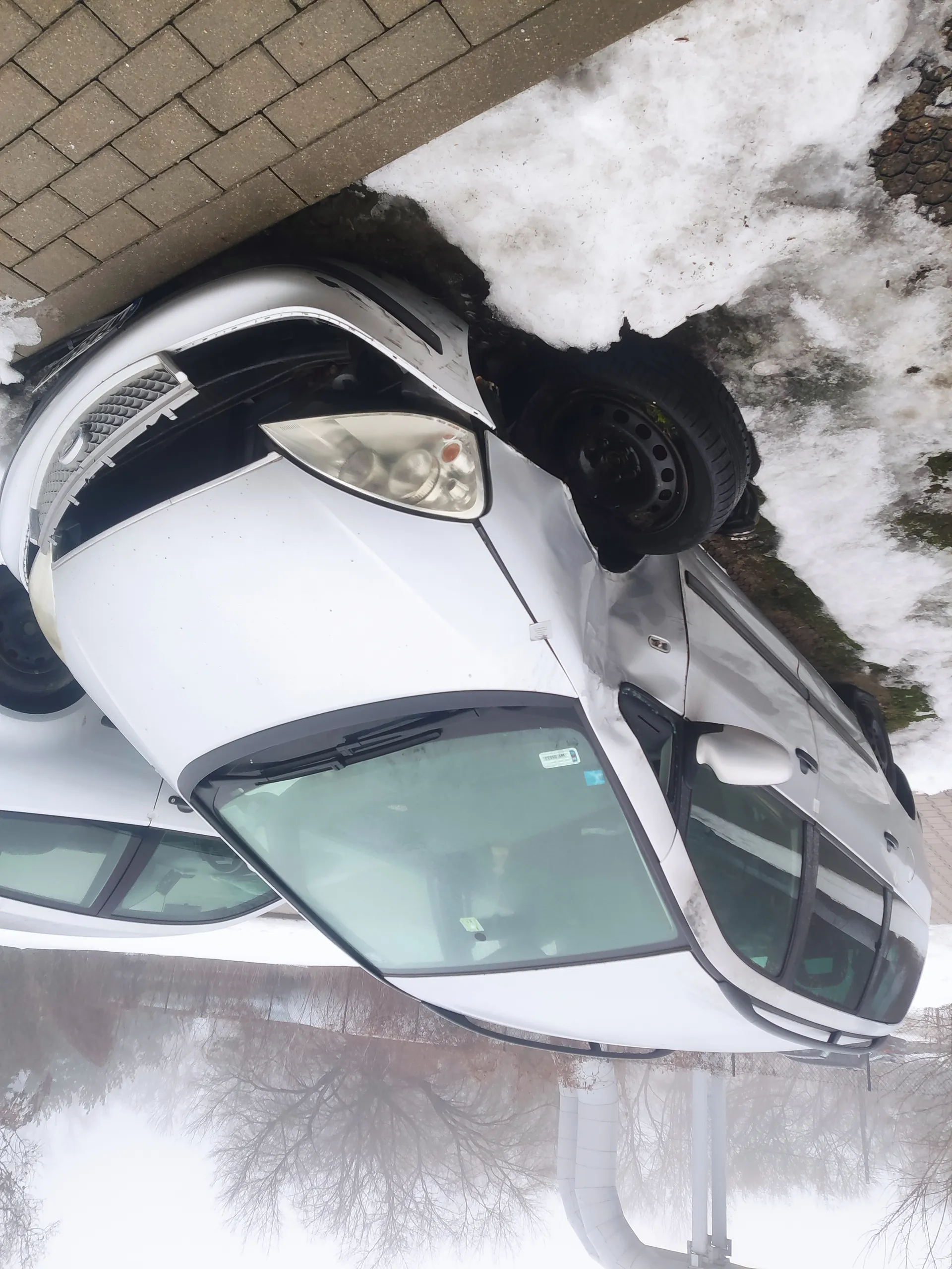 Biały samochód z uszkodzonym przodem, w tym zdeformowanym zderzakiem i uszkodzonym reflektorem, zaparkowany na śniegu obok chodnika z brązowych płytek. W tle drugi biały samochód, nagie drzewa i mgła. Na przedniej szybie widoczne są naklejki.