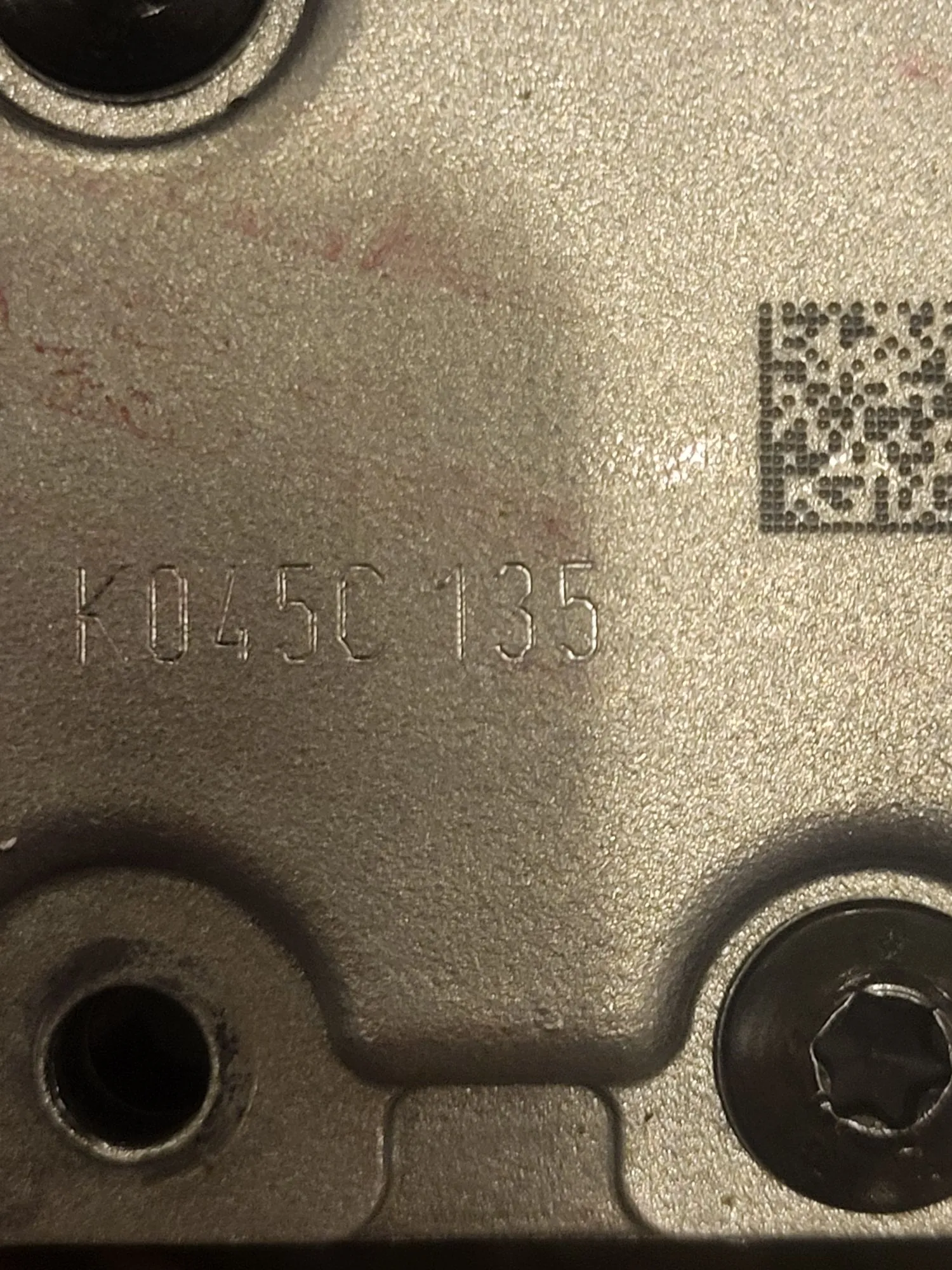 Zbliżenie na metalową, srebrną powierzchnię elementu mechanicznego. Na powierzchni znajduje się wytłoczony napis „K045C 135” oraz kod QR w prawym górnym rogu. W rogach widoczne są otwory montażowe, z czego jeden w prawym dolnym rogu jest wypełniony śrubą z łbem typu Torx.