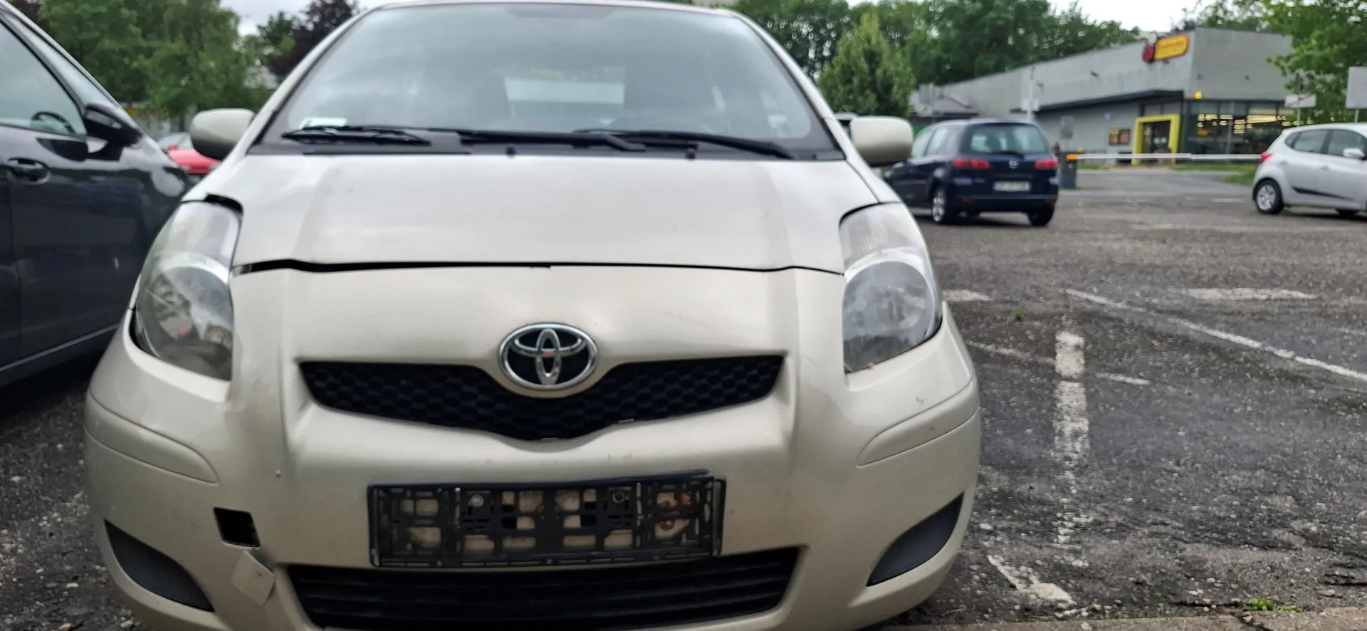Zdjęcie przodu jasnego samochodu marki Toyota zaparkowanego na asfaltowym parkingu. Pojazd ma widoczne uszkodzenia: szczelina między maską a zderzakiem oraz brak tablicy rejestracyjnej w uchwycie. W tle widoczne są inne zaparkowane samochody oraz budynek handlowy i drzewa.