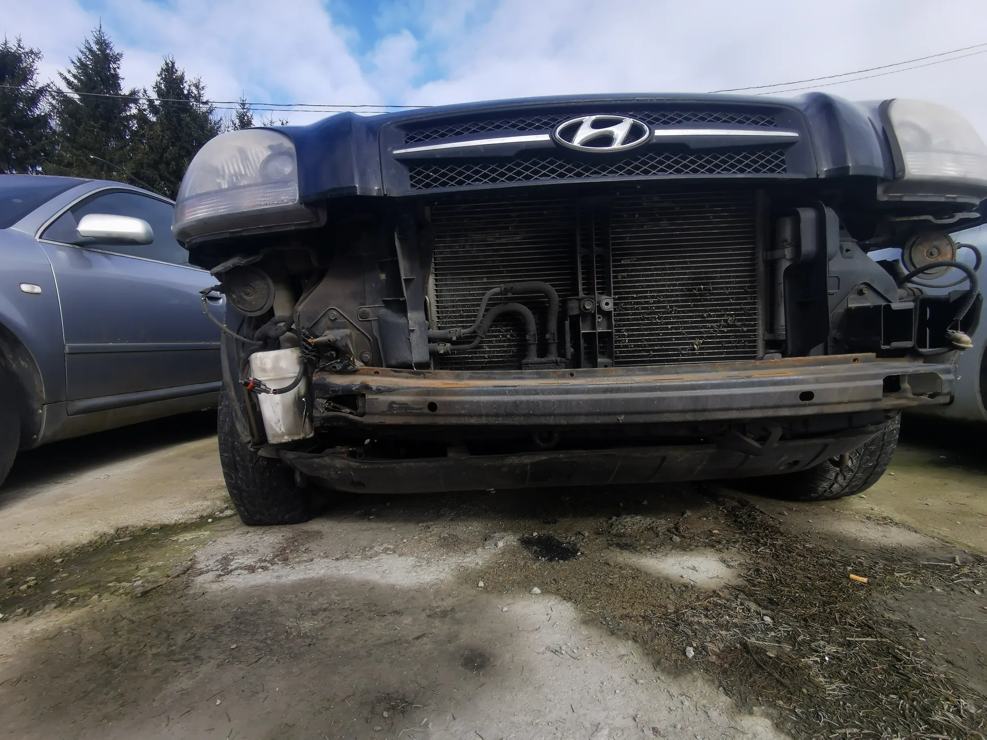 Przednia część uszkodzonego czarnego samochodu marki Hyundai z widocznym logo na atrapie chłodnicy. Brakuje przedniego zderzaka, widoczna jest chłodnica oraz elementy podwozia i przewody. Po lewej stronie częściowo widoczny szary samochód. Tło: drzewa, niebo z chmurami, betonowa powierzchnia z plamami i suchą trawą.
