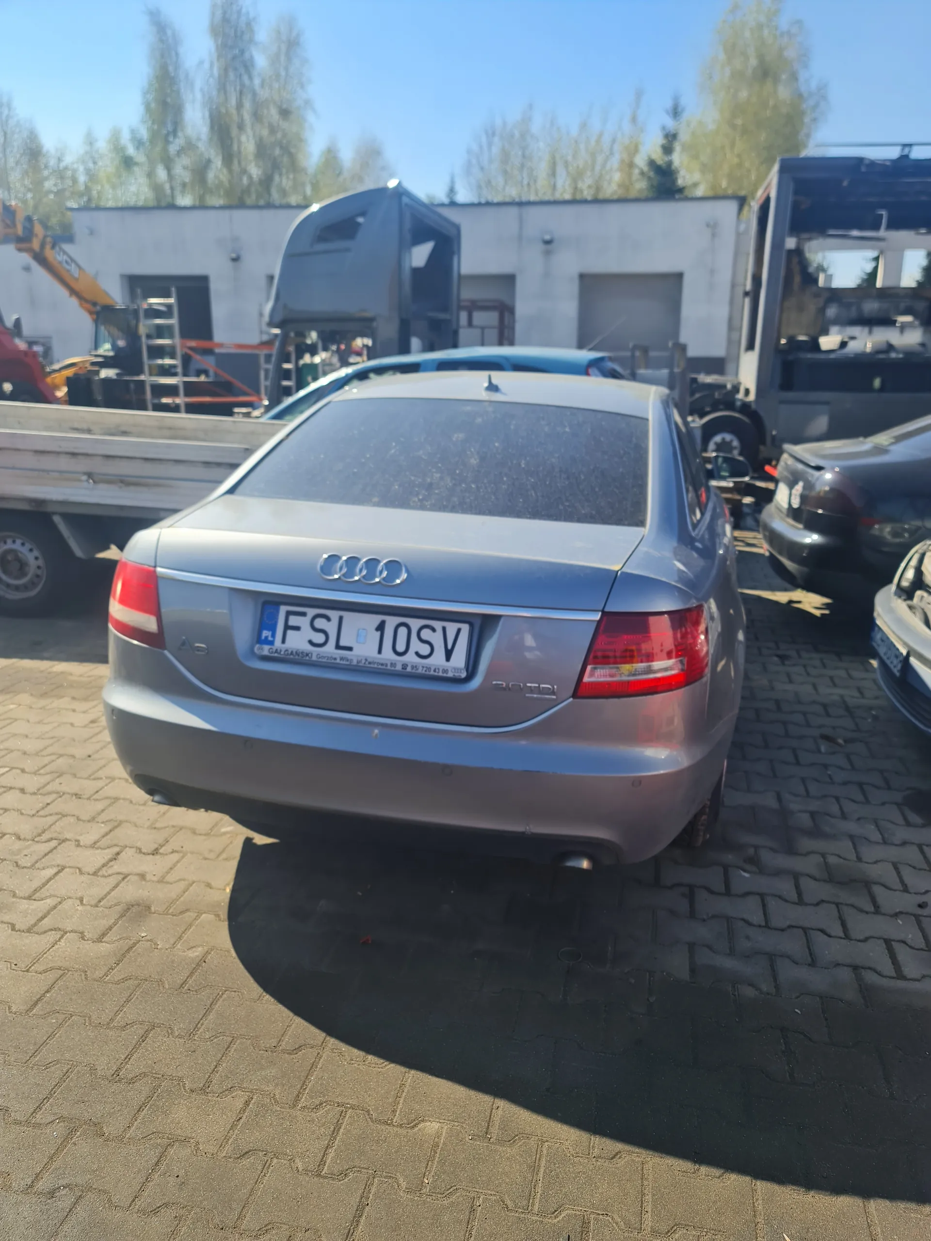 Srebrny samochód Audi A6 z tablicą rejestracyjną FSL 10SV zaparkowany na powierzchni z kostki brukowej. W tle widoczne są inne pojazdy, budynek z otwartymi przestrzeniami, maszyny budowlane oraz drzewa pod jasnym niebem. Tablica rejestracyjna zawiera napis PL oraz dodatkowe informacje w dolnej części.