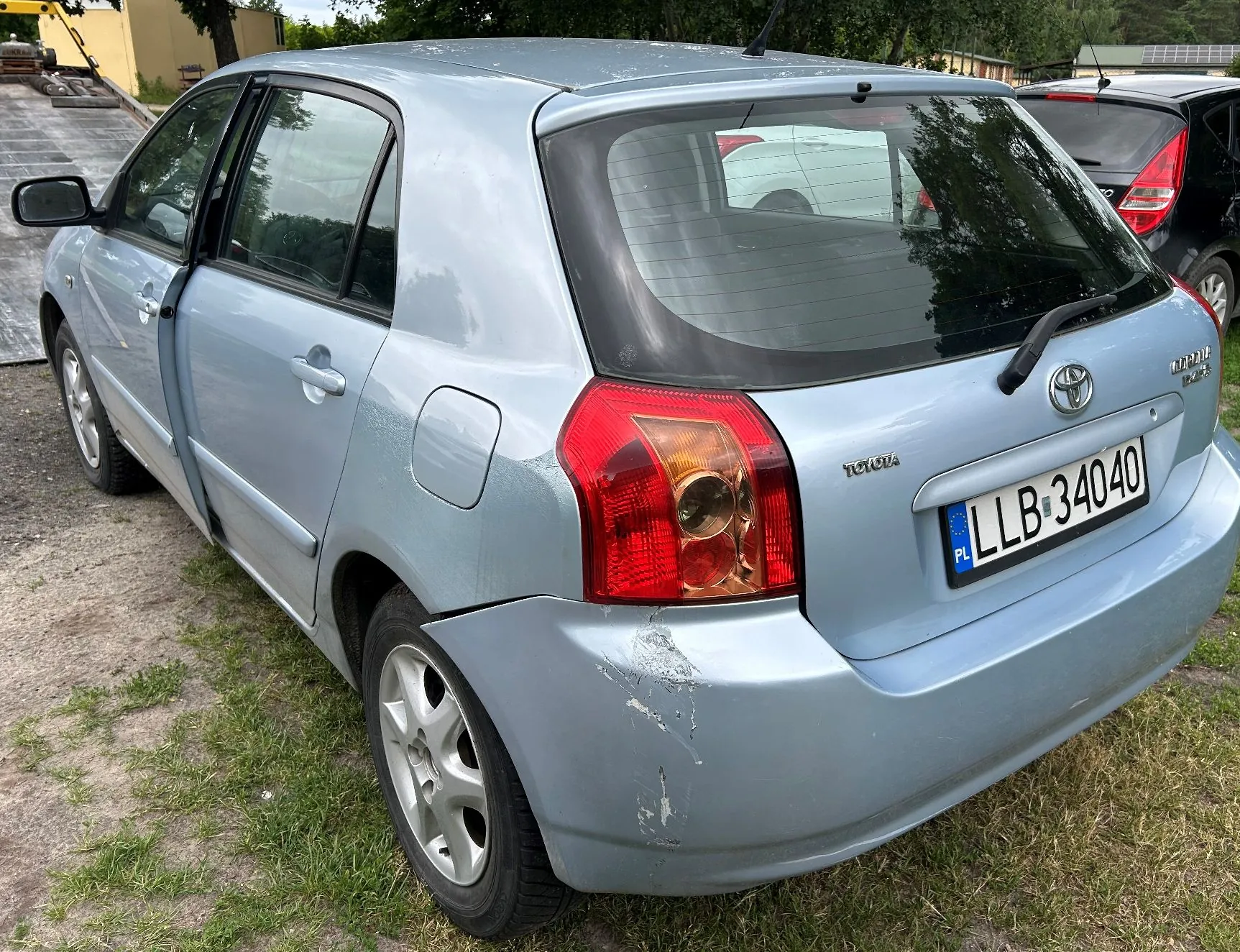Jasnoniebieski samochód marki Toyota Corolla w wersji hatchback, zaparkowany na trawie. Zdjęcie przedstawia tył i lewy bok pojazdu. Na tylnym zderzaku, po lewej stronie, widoczne są zarysowania i uszkodzenia lakieru. Samochód posiada polską tablicę rejestracyjną o numerze LLB 34040. W tle widoczny jest fragment innego, czarnego samochodu oraz zieleń drzew.