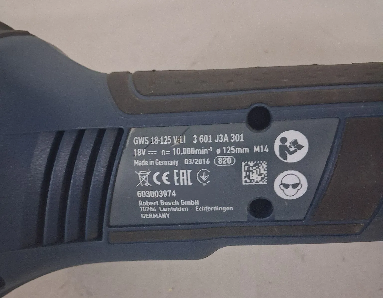 Zbliżenie na tabliczkę znamionową narzędzia marki Bosch. Na srebrnej etykiecie widnieją napisy: 'GWS 18-125 V-LI 3 601 J3A 301', '18V ⎓ n= 10.000min-1 ø 125mm M14', 'Made in Germany 03/2016 820'. Poniżej znajdują się symbole certyfikacji: CE, EAC oraz piktogramy dotyczące ochrony słuchu i oczu. Na dole widnieje numer '603003974' oraz dane producenta: 'Robert Bosch GmbH, 70764 Leinfelden-Echterdingen, GERMANY'.