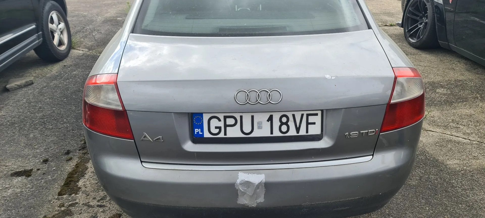 Tylny widok srebrnego samochodu marki Audi A4. Na klapie bagażnika znajduje się logo Audi (cztery splecione pierścienie), tablica rejestracyjna o numerze GPU 18VF z oznaczeniem PL oraz oznaczenia modelu 'A4' po lewej i silnika '1.9 TDI' po prawej stronie. Na zderzaku widoczny jest biały fragment taśmy lub kleju.