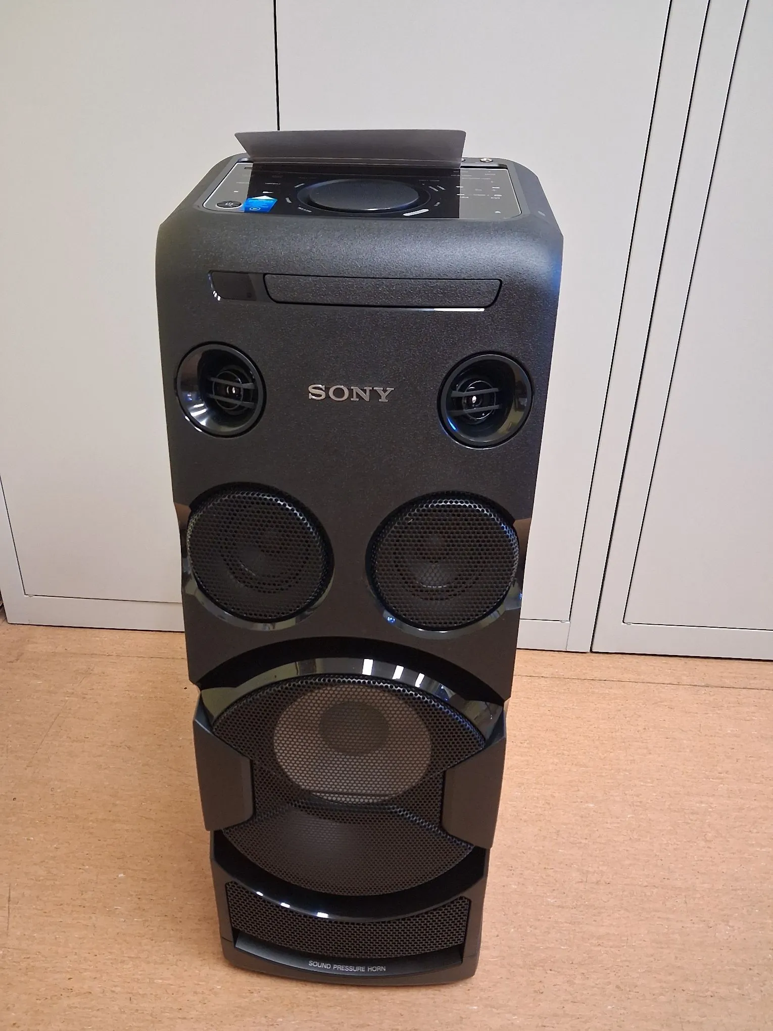 Czarny, wysoki głośnik wieżowy marki Sony. Urządzenie posiada logo SONY na przednim panelu, dwa mniejsze głośniki w górnej części, dwa średniej wielkości głośniki w środkowej sekcji oraz jeden duży głośnik basowy na dole. Na górze znajduje się panel sterowania z przyciskami i pokrętłem. Na samym dole widnieje napis SOUND PRESSURE HCPN.