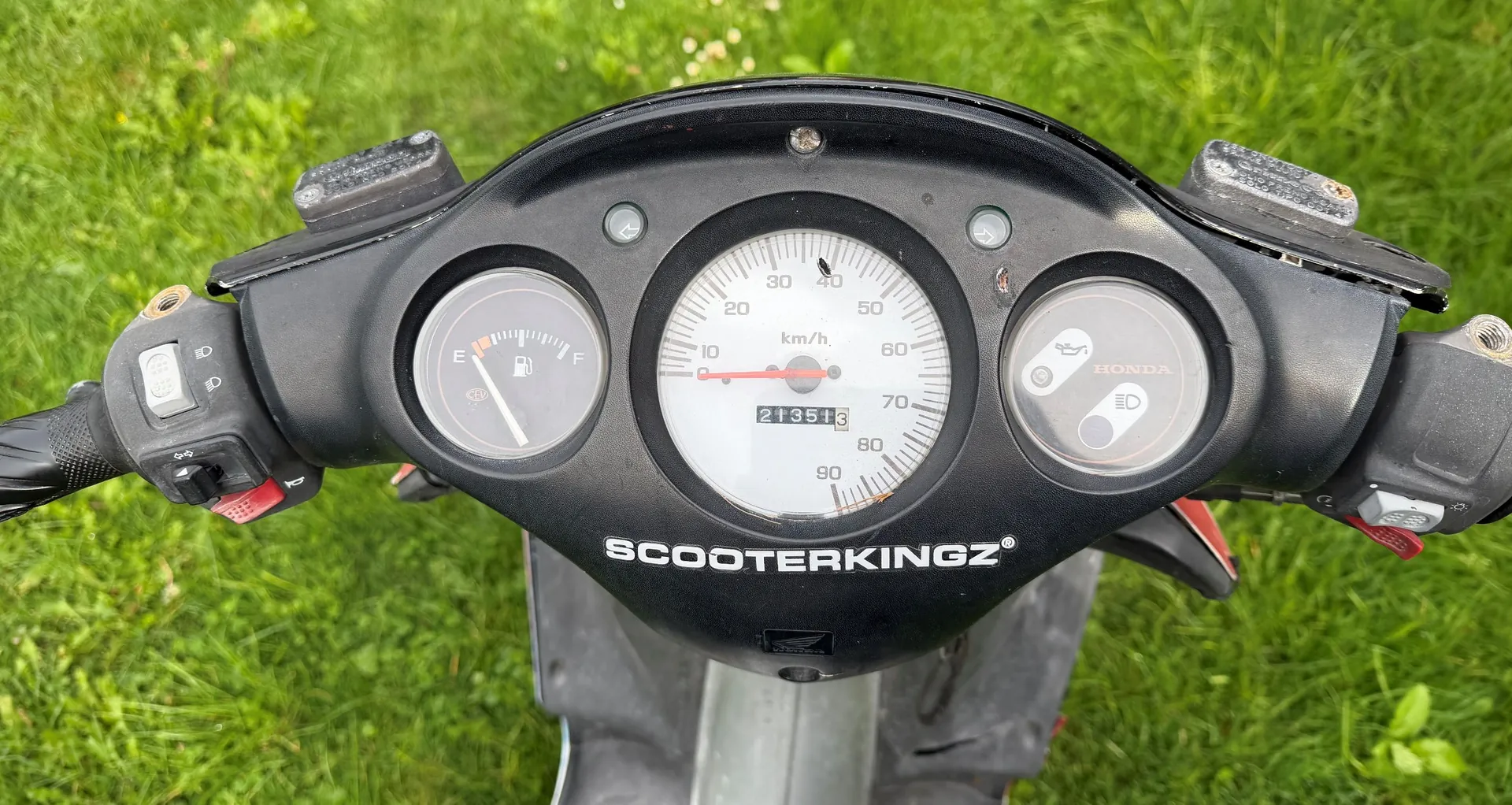 Widok z perspektywy kierowcy na deskę rozdzielczą czarnego skutera marki Honda. W centrum znajduje się analogowy prędkościomierz z podziałką do 90 km/h i licznikiem przebiegu wskazującym 21351. Po lewej stronie znajduje się wskaźnik poziomu paliwa z oznaczeniami 'E' i 'F'. Po prawej stronie widnieje logo HONDA oraz kontrolka świateł. Poniżej zegarów znajduje się biały napis 'SCOOTERKINGZ®'. Skuter stoi na tle zielonej trawy.