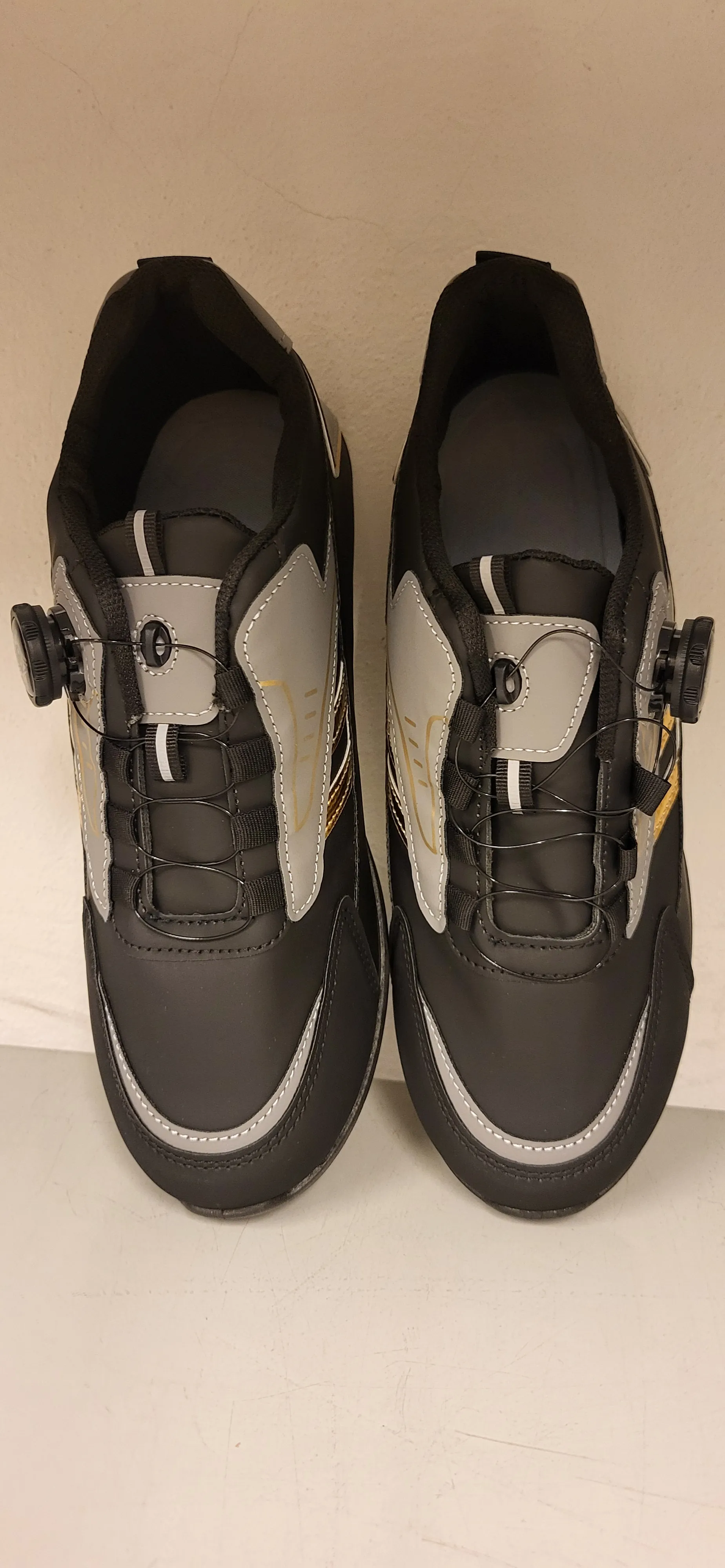 Dwa czarne buty rowerowe z szarymi i złotymi akcentami, z zamkami typu kołnierz i sznurowaniem, z białymi przeszyciami. Buty ułożone są na jasnej powierzchni.