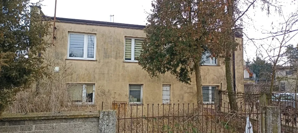 Zdjęcie starego, dwukondygnacyjnego budynku o żółtawej, zniszczonej elewacji. Na piętrze widoczne są trzy okna, z czego jedno ma białe żaluzje. Na parterze znajdują się trzy okna oraz jedne zamknięte białe drzwi. Budynek jest częściowo zasłonięty przez wysokie, zielone drzewa i krzewy. Przed domem znajduje się metalowy płot z pionowymi prętami oraz niski murek z betonowych bloczków.