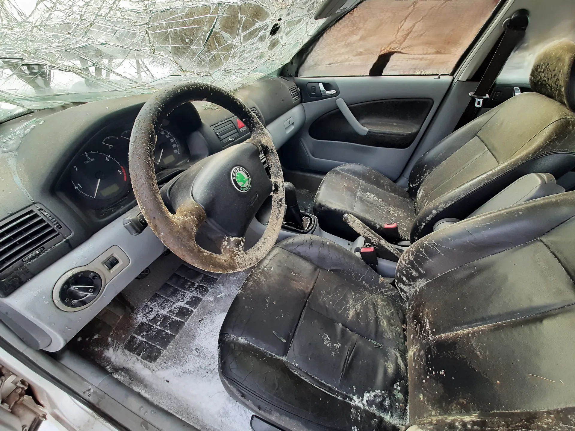 Wnętrze samochodu marki Skoda z uszkodzonym, pękniętym przednim szybą. Widoczna kierownica z logo Skoda, zniszczone czarne fotele pokryte brudem i śniegiem, deska rozdzielcza z licznikami oraz szare panele drzwiowe. Wnętrze wydaje się zapuszczone, z widocznymi śladami zużycia i zanieczyszczeń.
