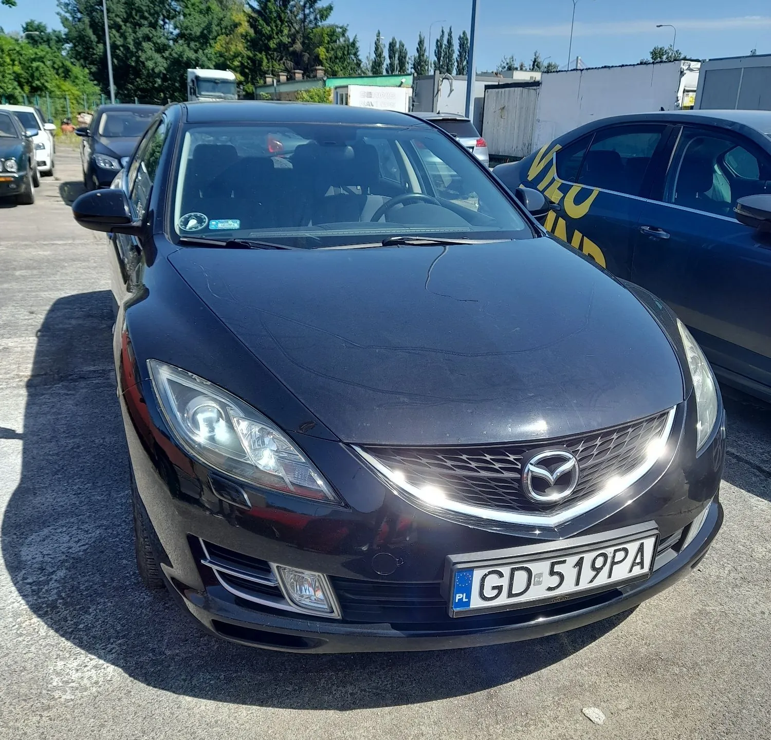 Czarny samochód marki Mazda zaparkowany na zewnątrz. Widoczny jest przód pojazdu z logo Mazdy na grillu oraz polska tablica rejestracyjna o numerze GD 519PA. W tle znajdują się inne samochody, budynki przemysłowe i drzewa pod jasnym, niebem.