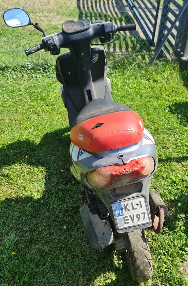 Widok z tyłu na skuter zaparkowany na zielonej trawie. Skuter ma szary kolor z czerwonym siedzeniem, na którym widnieje napis '700'. Tylna lampa jest uszkodzona i pęknięta. Pojazd posiada polską tablicę rejestracyjną o numerze KL I EV97. W tle widoczny jest metalowy płot i trawnik.