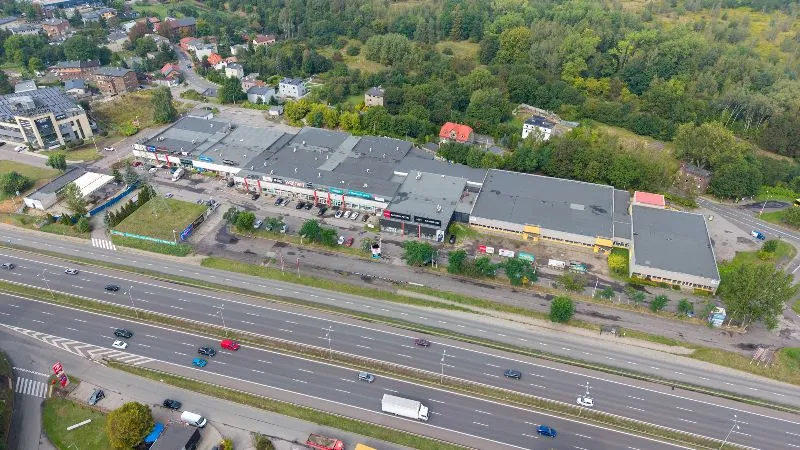 Widok z lotu ptaka na kompleks handlowo-usługowy z dużymi, płaskimi dachami, otoczony parkingami i zielenią. Przed budynkami przebiega wielopasmowa droga szybkiego ruchu z kilkoma jadącymi samochodami i ciężarówką. W tle widoczne są domy jednorodzinne oraz gęsty las.
