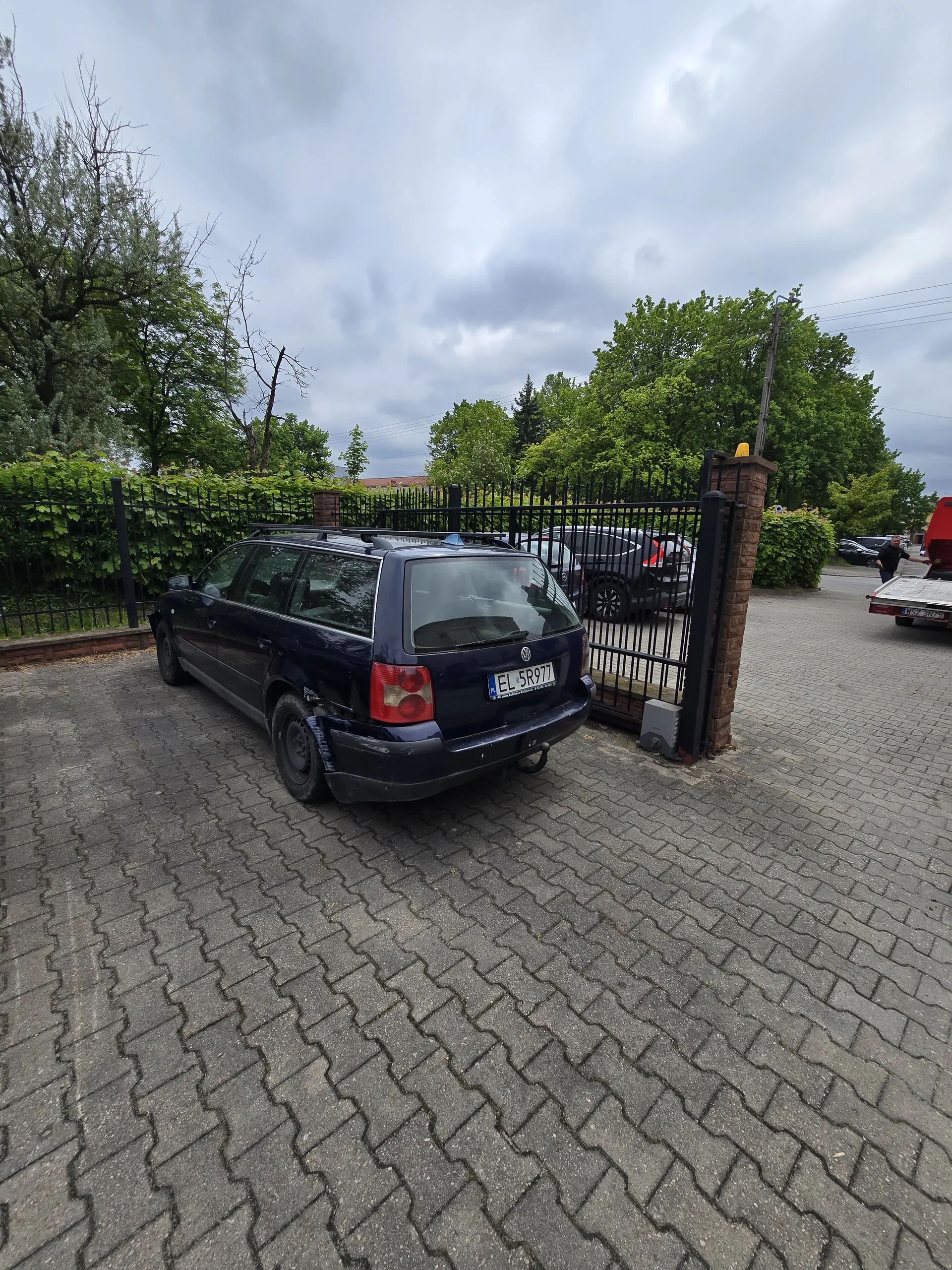 Ciemnoniebieski samochód zaparkowany na brukowanej powierzchni. Widoczna tablica rejestracyjna EL 5R971. Za czarną metalową bramą z ceglanymi filarami znajduje się inny samochód. W tle drzewa, zielone krzewy i zachmurzone niebo. Na drodze w oddali czerwony pojazd.