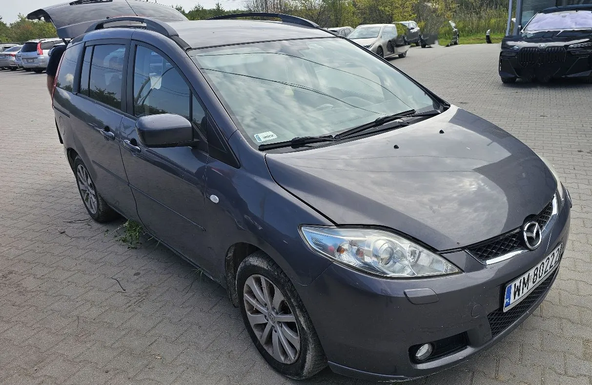 Srebrno-szary samochód marki Mazda w nadwoziu kombi, zaparkowany na kostce brukowej. Pojazd jest widoczny z przodu i z boku. Na przednim zderzaku znajduje się tablica rejestracyjna o numerze WM 80227. W tle widoczne są inne zaparkowane samochody oraz fragment budynku.