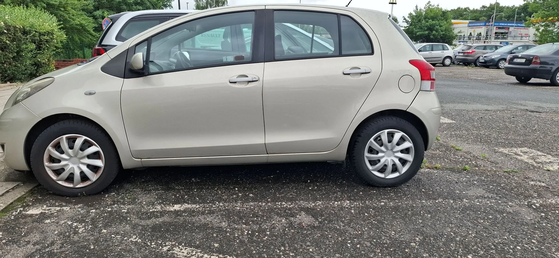 Profil boczny beżowego samochodu osobowego typu hatchback zaparkowanego na asfaltowym placu. W tle widoczne są inne zaparkowane samochody, zielone krzewy oraz fragmenty budynków handlowych.