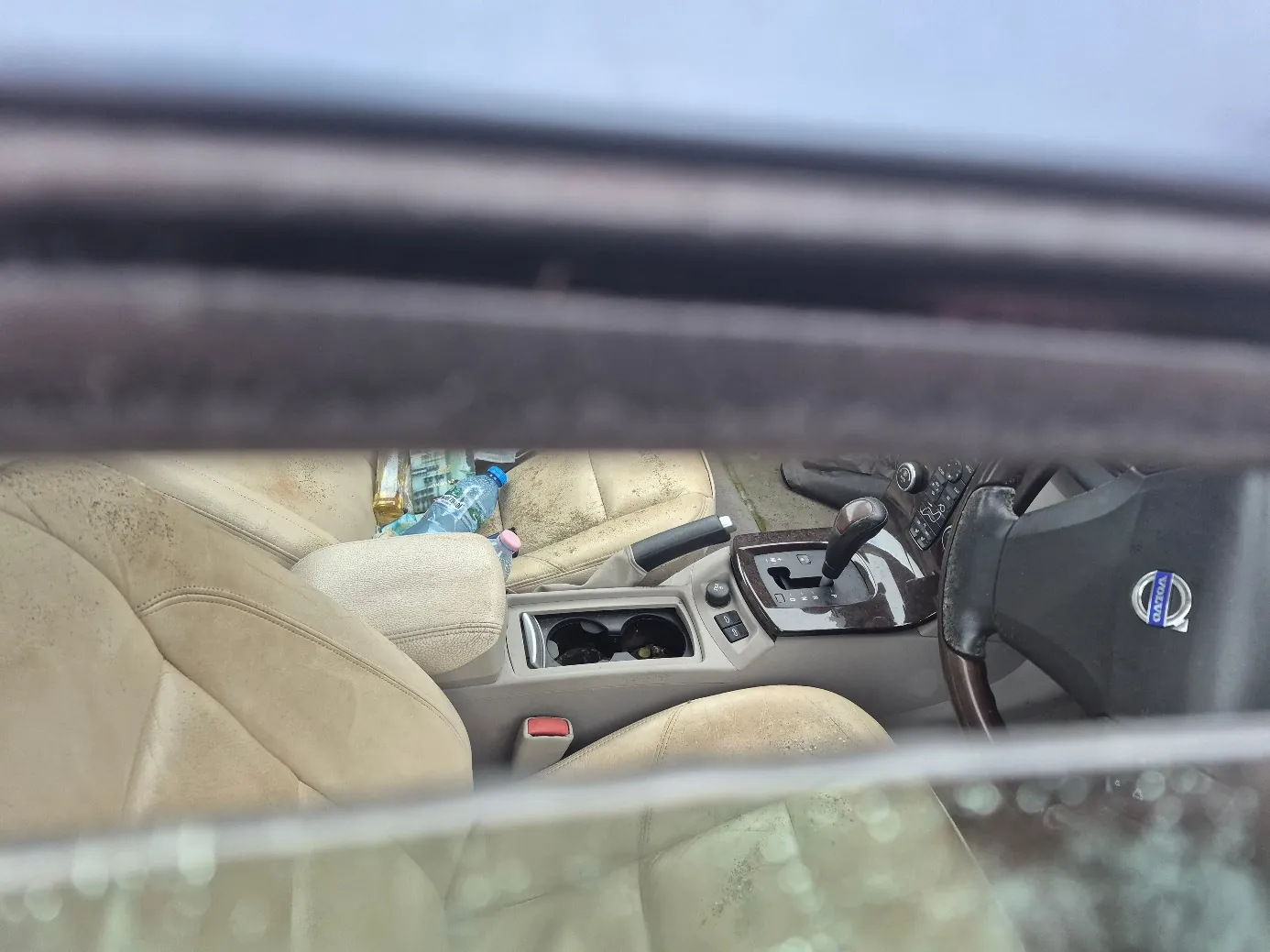 Wnętrze samochodu marki Volvo widziane przez przednią szybę. Beżowe, zabrudzone fotele, kierownica z logo Volvo, centralna konsoła z dźwignią biegów i uchwytem na napoje. Na fotelu pasażera leżą plastikowa butelka wody i inne przedmioty. Szyba przednia ma ślady zabrudzeń.