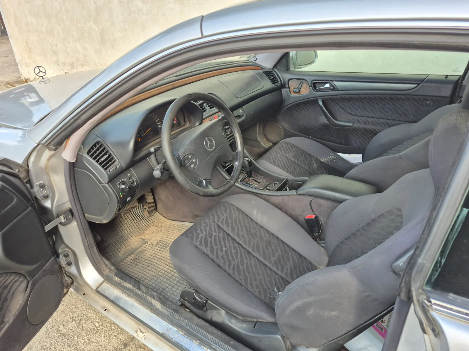 Wnętrze samochodu marki Mercedes-Benz z otwartą przednią lewą drzwiami. Widoczne są czarne siedzenia z wzorzystą tapicerką, kierownica z logo marki, deska rozdzielcza z drewnianą obwódką, zanieczyszczone dywaniki podłogowe oraz fragment srebrnego nadwozia. W tle betonowa ściana.