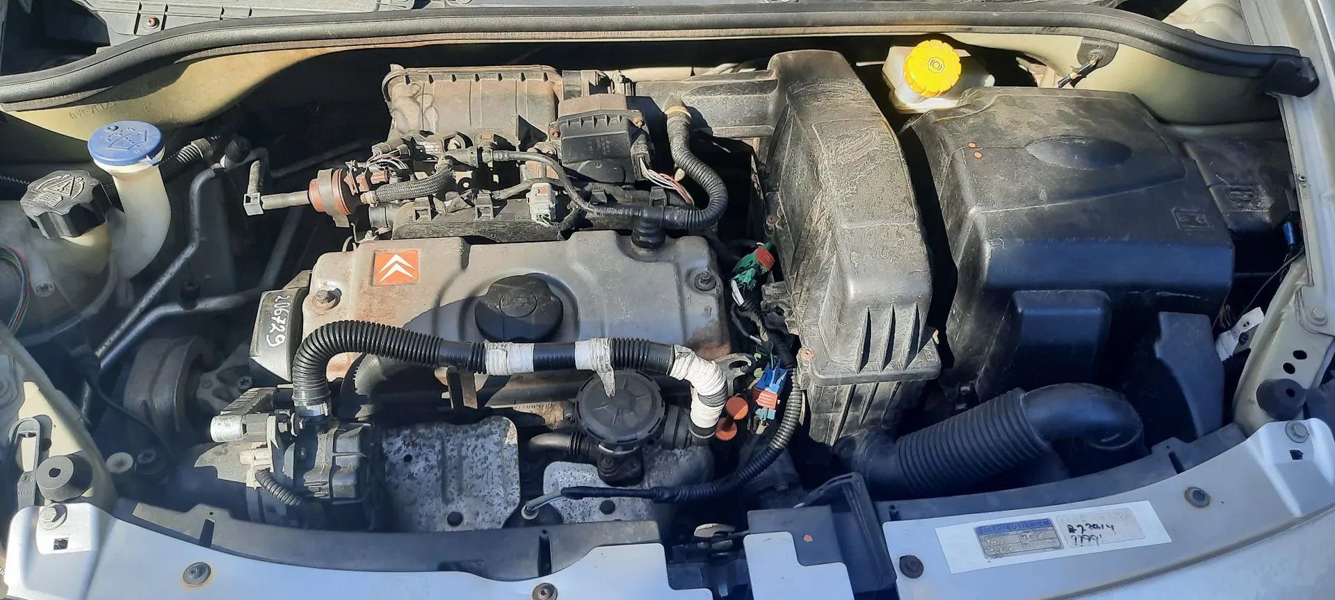Widok z góry na komorę silnika samochodu marki Citroën. Widoczny jest silnik z logo marki na plastikowej osłonie, zbiornik płynu chłodniczego z niebieskim korkiem po lewej stronie, czarna obudowa filtra powietrza po prawej oraz różnorodne przewody, wiązki elektryczne i elementy mechaniczne. W dolnej części zdjęcia widoczna jest biała rama nadwozia z naklejką techniczną.
