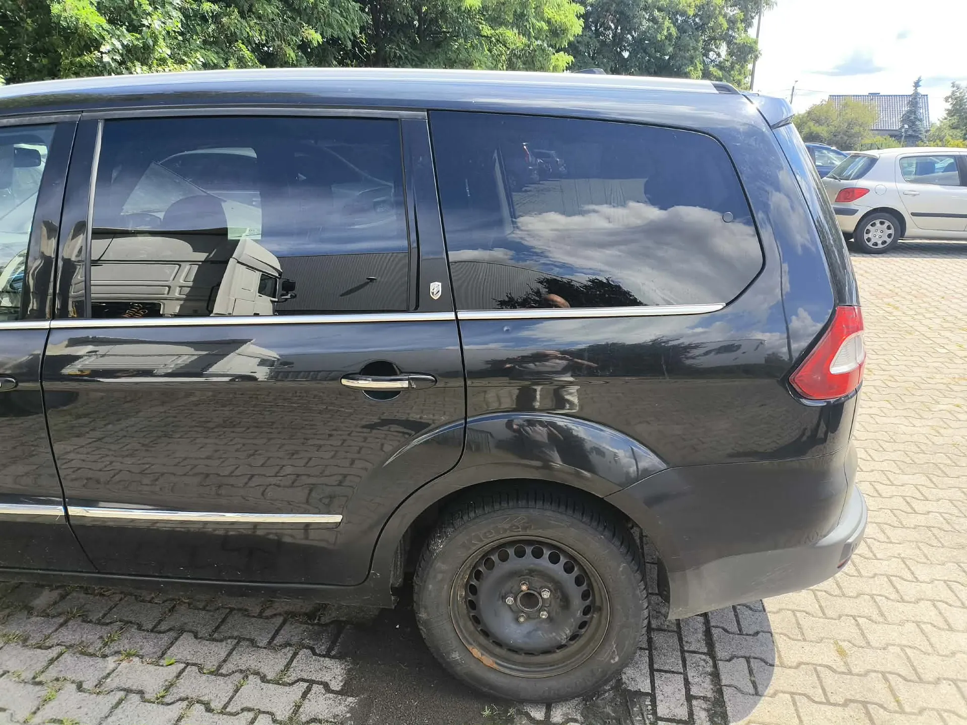 Boczny widok czarnego samochodu typu minivan zaparkowanego na kostce brukowej. Pojazd ma przyciemniane szyby i srebrne listwy ozdobne wzdłuż dolnej części drzwi oraz pod oknami. W tle widoczne są zielone drzewa oraz inny, biały samochód zaparkowany obok.