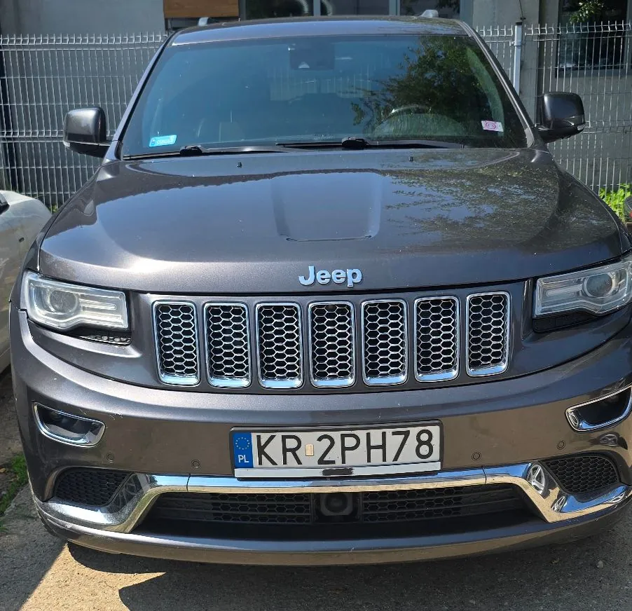 Widok z przodu na brązowy samochód marki Jeep. Pojazd posiada charakterystyczny grill z siedmioma pionowymi szczelinami oraz tablicę rejestracyjną o numerze KR 2PH78. Samochód stoi zaparkowany przed metalowym ogrodzeniem.