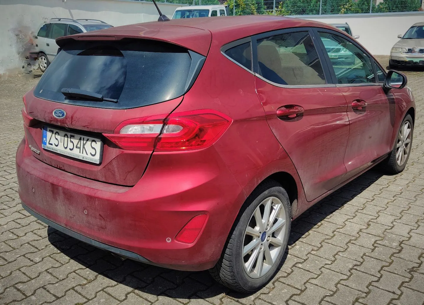 Czerwony samochód marki Ford Fiesta zaparkowany na kostce brukowej. Widok z tyłu i z boku prawego. Pojazd posiada polską tablicę rejestracyjną o numerze ZS 054KS. W tle widoczne są inne samochody oraz biały mur.