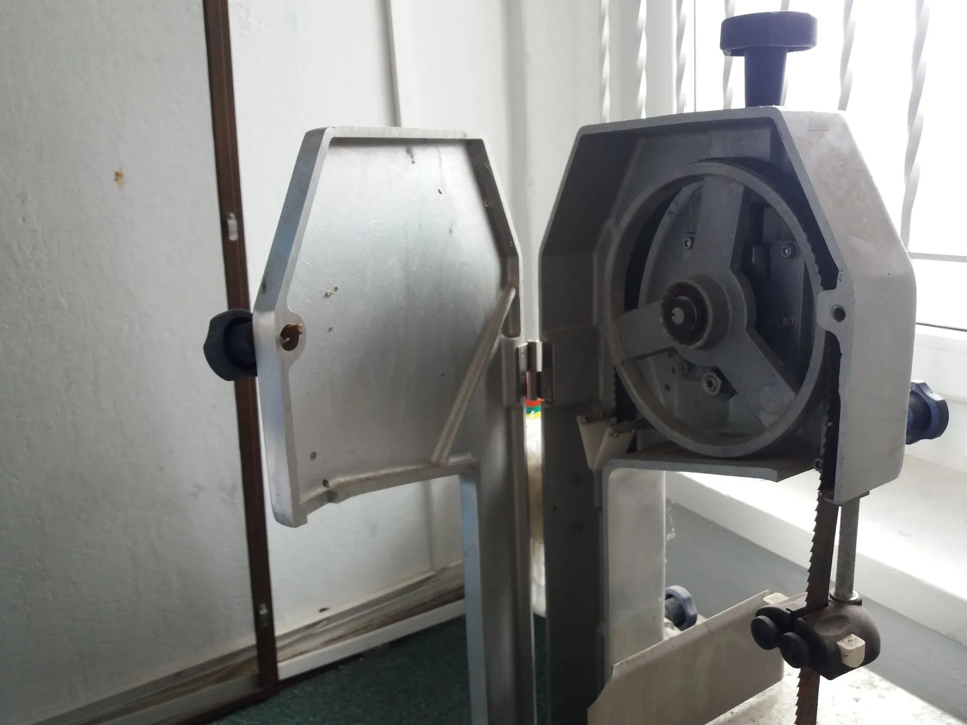 Zbliżenie na metalową maszynę przemysłową, prawdopodobnie piłę taśmową. Widoczny jest mechanizm z kołem pasowym, napędem taśmowym oraz aluminiową obudową i elementami konstrukcyjnymi. Urządzenie znajduje się w jasnym pomieszczeniu przy oknie.