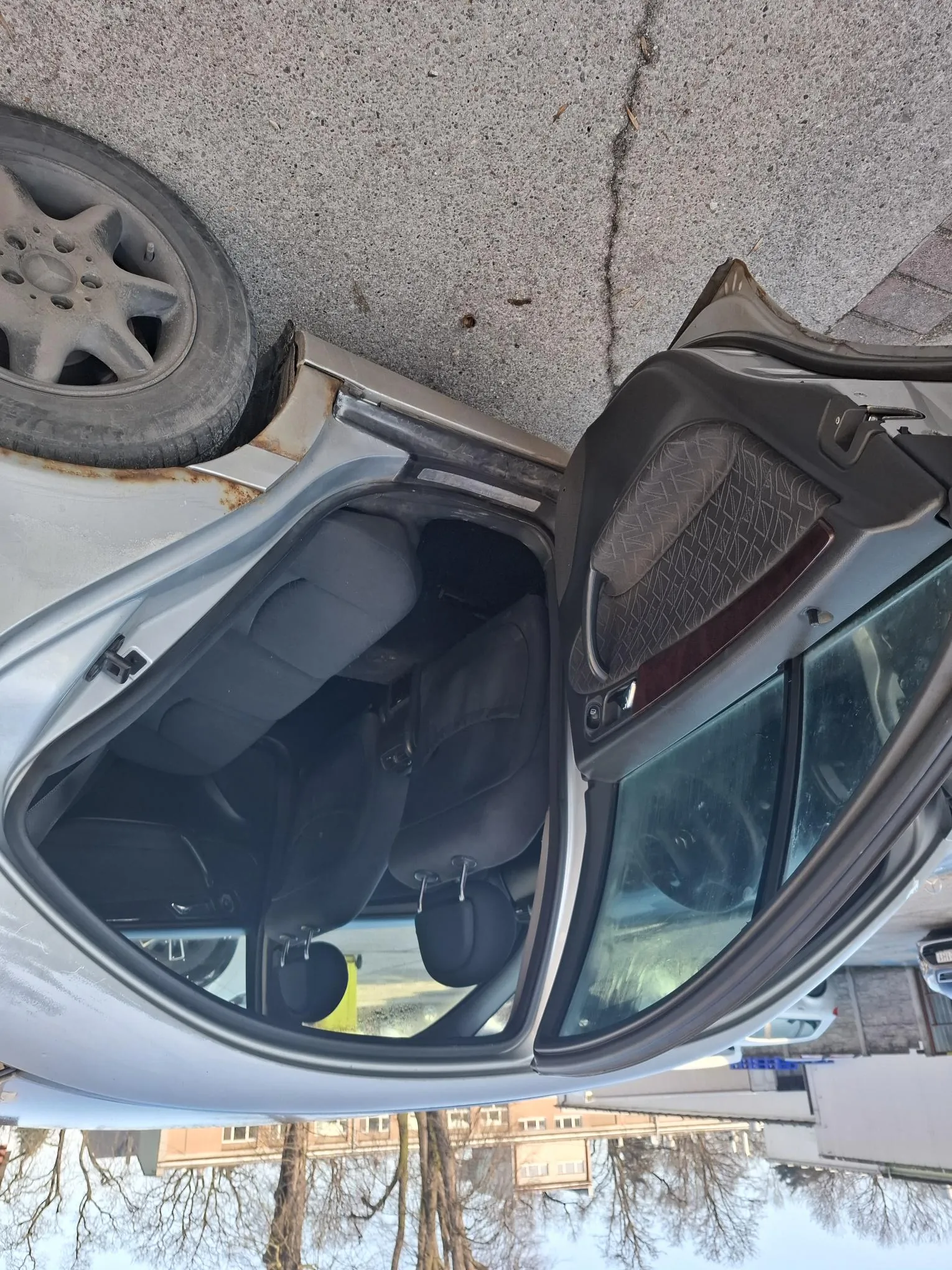 Srebrny samochód z otwartą przodem kierowcy. Widoczne czarne siedzenia, zardzewiałe miejsce koło koła oraz tło z budynkami, drzewami bez liści i nawierzchnią asfaltową.