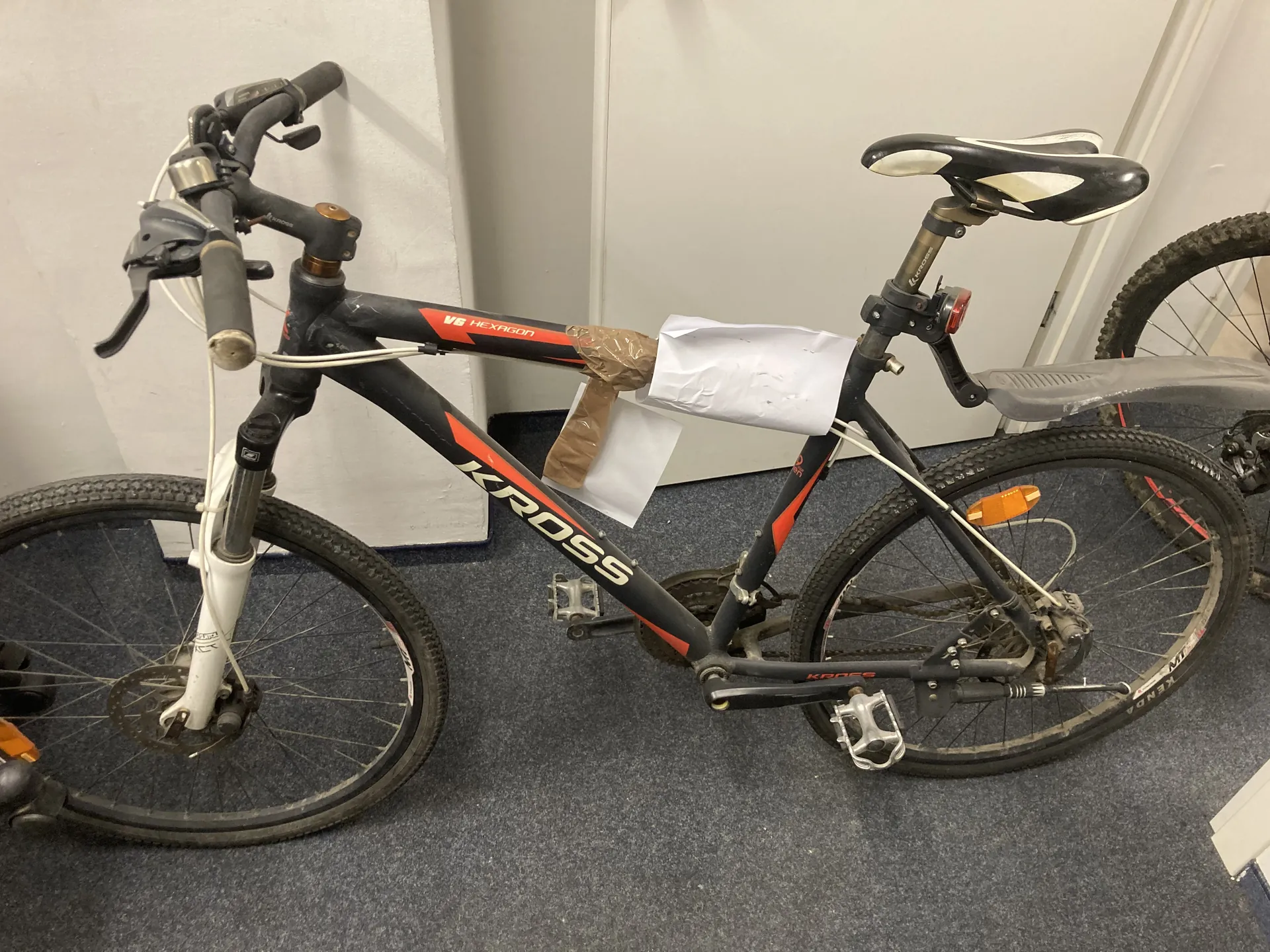 Rower marki KROSS o czarno-czerwonym kolorze z widocznym napisem 'KROSS' na ramie oraz 'V6 HEXAGON' na górnej części ramy. Rower posiada czarno-białe siedzisko, przedni widelec amortyzacyjny, hamulce tarczowe i pomarańczowe reflektory. Na ramie zamocowano biały arkusz papieru oraz brązową taśmę. Rower stoi na szarym dywanie przy białej ścianie. W tle częściowo widoczne koło innego roweru. Na oponie tylnego koła widoczny napis 'KENDA'.