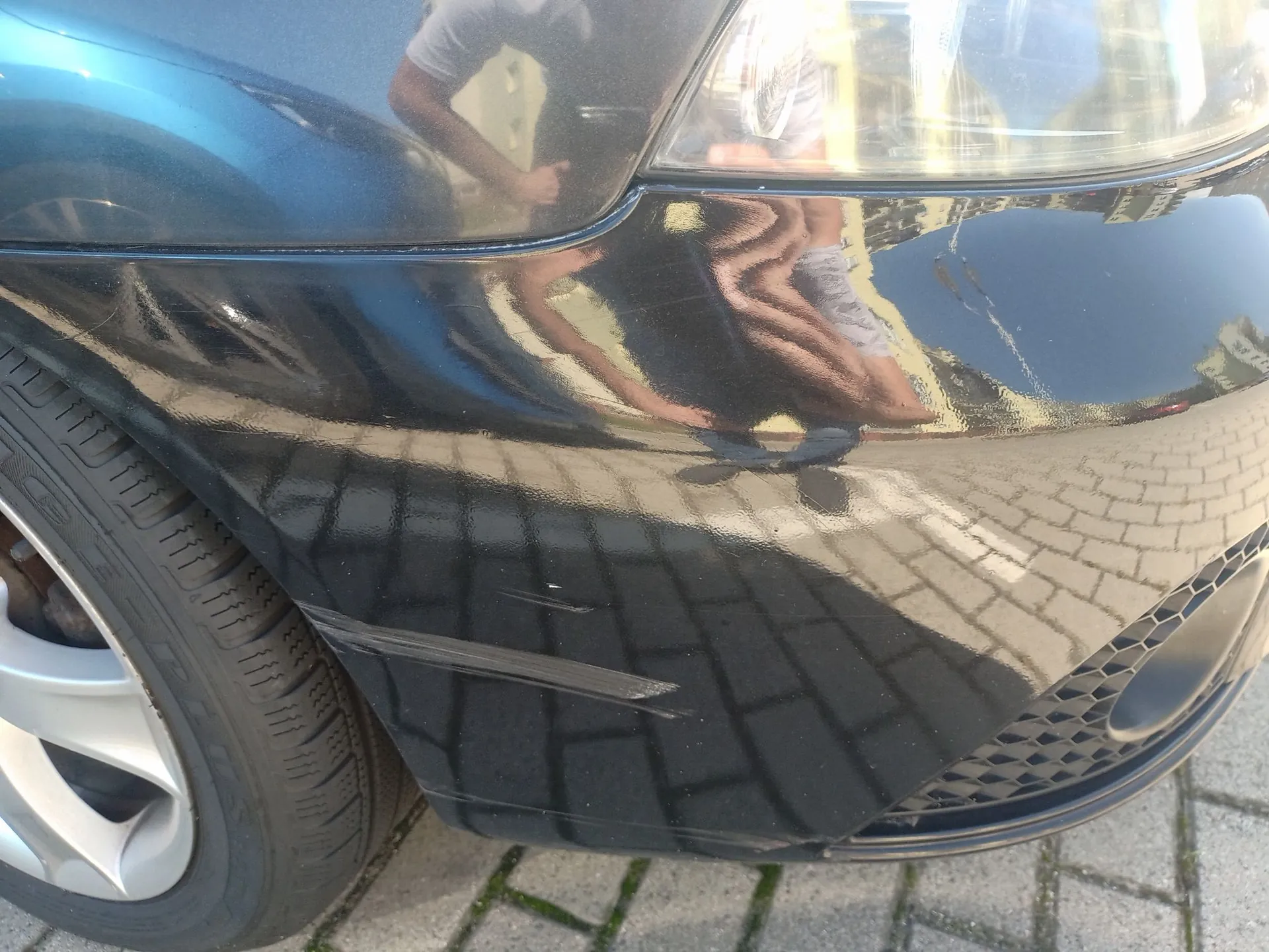 Zbliżenie na przedni prawy narożnik ciemnoniebieskiego samochodu. Na zderzaku widoczne są zarysowania oraz wgniecenie. W lakierze odbija się sylwetka osoby robiącej zdjęcie oraz kostka brukowa. W lewej części kadru widoczny jest fragment koła z felgą aluminiową i oponą.