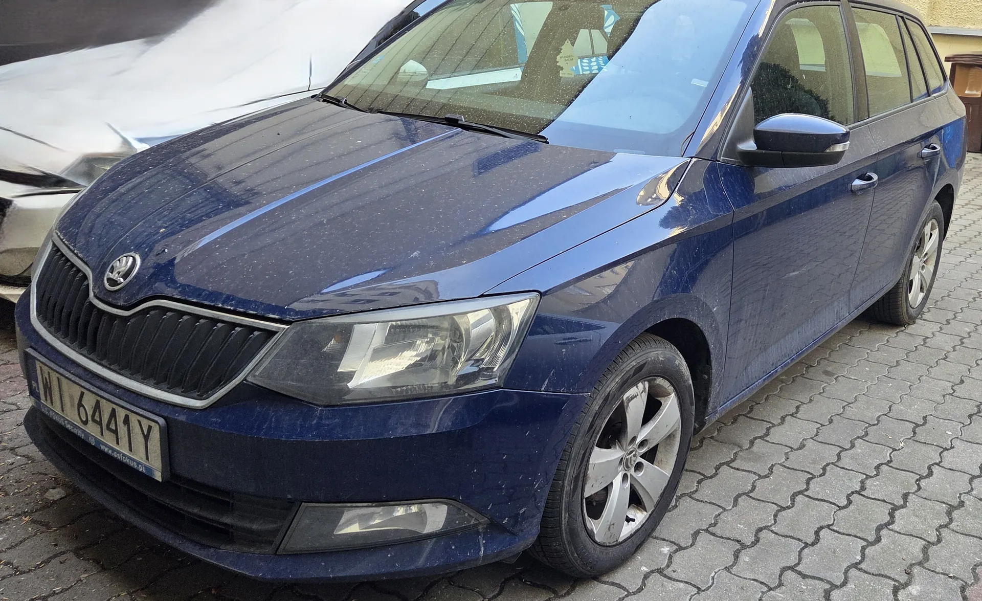 Niebieski samochód marki Skoda, model kombi, zaparkowany na brukowanej powierzchni. Widoczna tablica rejestracyjna z numerem WI 6441Y. Samochód jest nieco zabrudzony. W tle częściowo widoczny srebrny samochód.