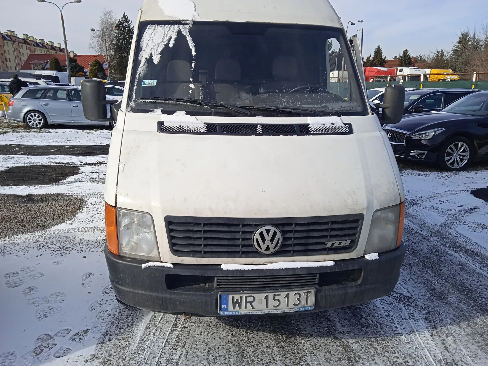 Biały samochód dostawczy Volkswagen TDI z przodu, z widocznym logo VW i tablicą rejestracyjną WR 1513T. Na masce, przedniej szybie i dachu leży śnieg. W tle parkujące samochody (srebrny kombi i czarny sedan), budynki mieszkalne, drzewa oraz śnieżna nawierzchnia z odciskami stóp. Na przedniej szybie widoczny nieczytelny naklejony dokument.