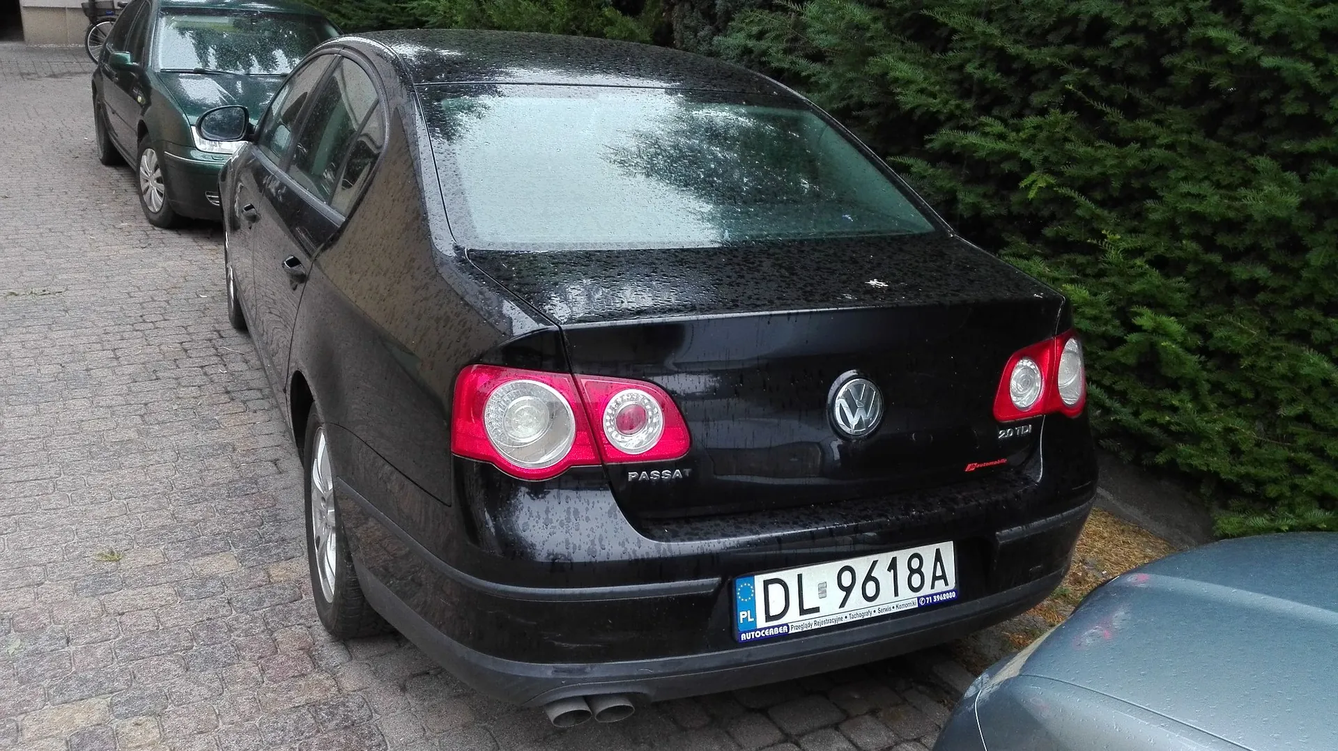 Czarny samochód marki Volkswagen Passat zaparkowany na kostce brukowej. Widok z tyłu i z boku. Samochód jest mokry od deszczu. Na klapie bagażnika widnieją napisy 'PASSAT' oraz '2.0 TDI'. Tablica rejestracyjna ma numer 'DL 9618A'. W tle widoczny jest fragment innego ciemnego samochodu oraz zielony żywopłot.