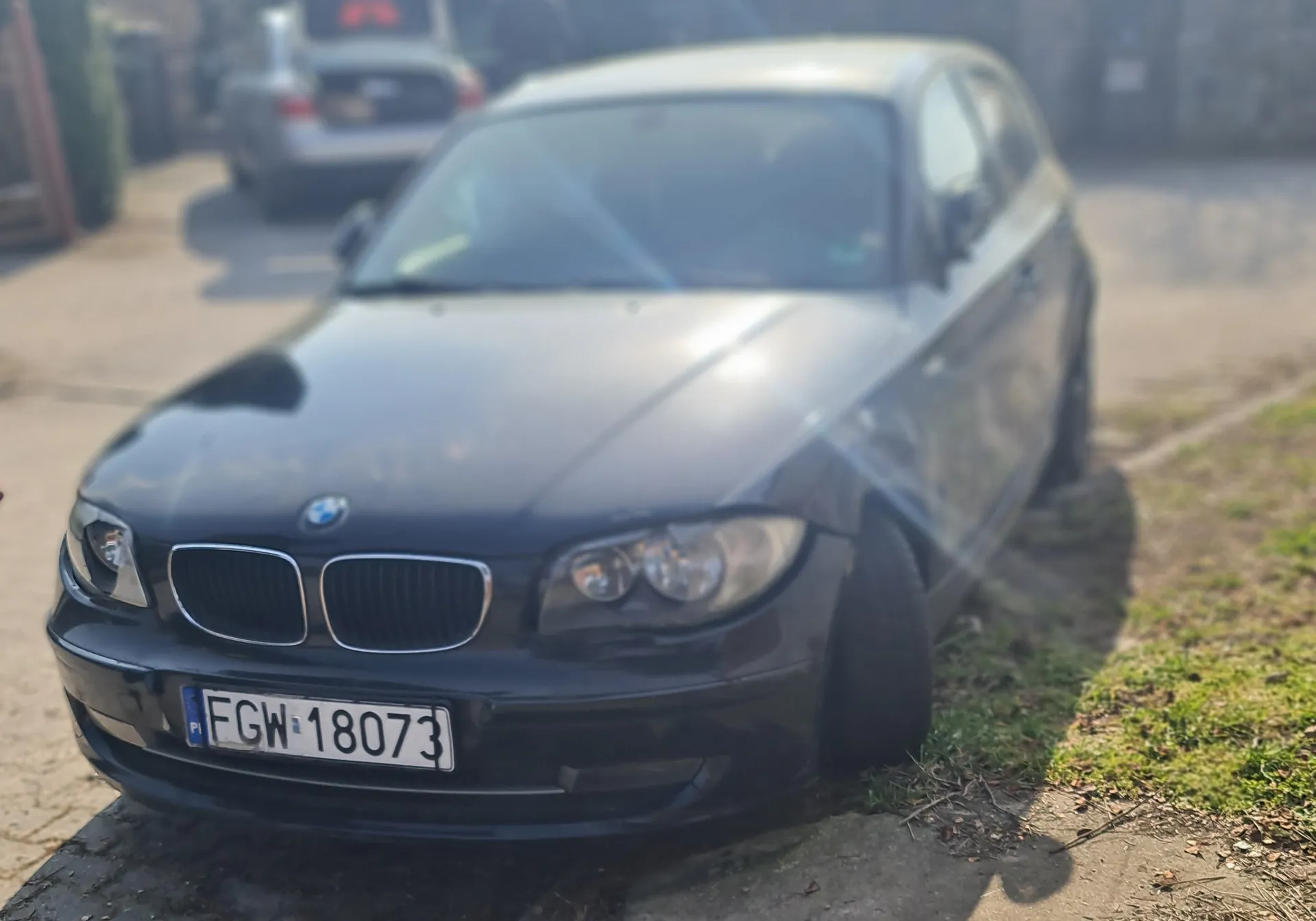 Czarny samochód marki BMW zaparkowany na poboczu drogi. Widoczna tablica rejestracyjna z białym tłem i czarnym napisem FGW 18073 oraz niebieskim pasem z oznaczeniem PL po lewej stronie. W tle drugi samochód, chodnik i fragment trawnika. Obraz jest nieco rozmyty.