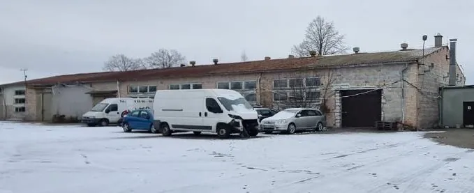 Zdjęcie przedstawia zimowy krajobraz z budynkiem przemysłowym o długim, jednopiętrowym kształcie z ceglanymi ścianami i brązowym dachem. Przed budynkiem, na śnieżnej powierzchni, zaparkowane są pojazdy: biała furgonetka z przodu, niebieski samochód osobowy, srebrny samochód osobowy oraz druga biała furgonetka w tle. W tle widoczne są bezlistne drzewa i pochmurne niebo. Budynek posiada duże okna, bramę garażową oraz kilka kominów na dachu.