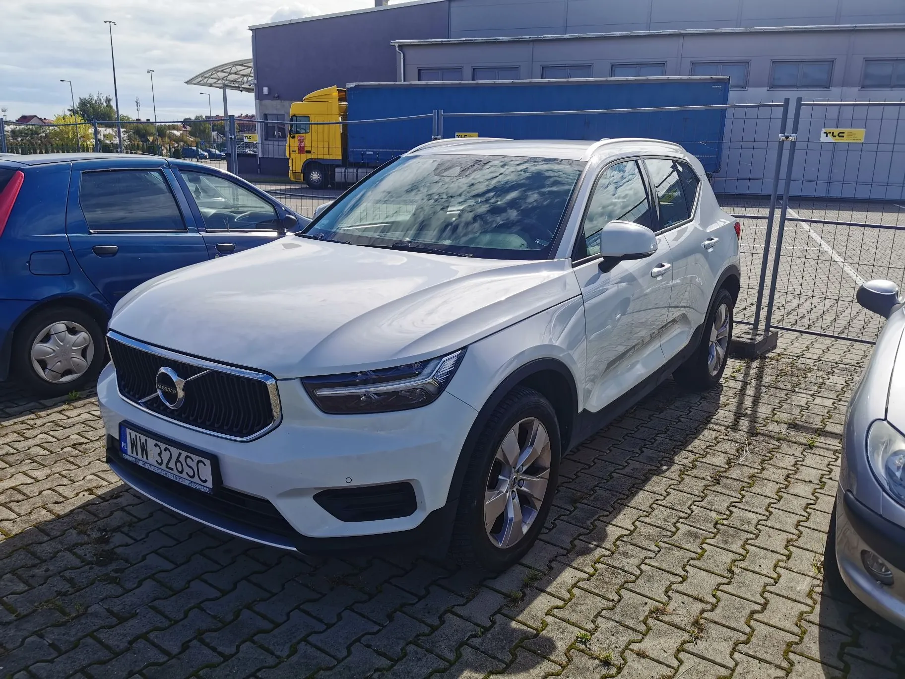 Biały samochód marki Volvo XC40 zaparkowany na kostce brukowej. Pojazd ma polską tablicę rejestracyjną o numerze WW 326SC. Po lewej stronie widoczny jest fragment niebieskiego samochodu, a po prawej fragment srebrnego auta. W tle znajduje się niebieska naczepa ciężarówki oraz szary budynek przemysłowy.