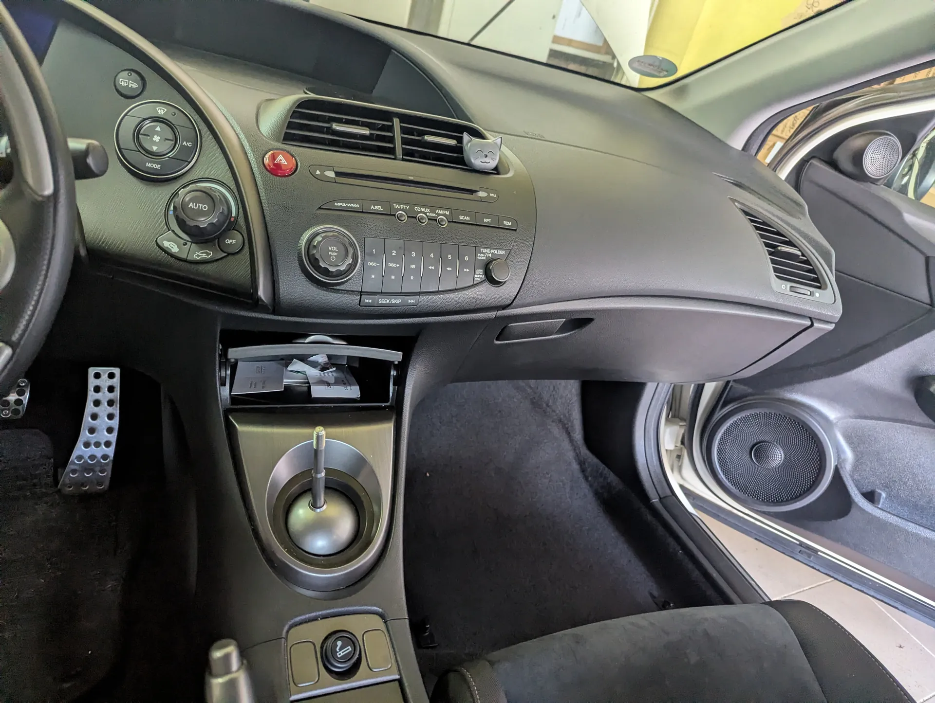 Wnętrze samochodu z widoczną deską rozdzielczą, kierownicą po lewej stronie, przyciskami klimatyzacji z napisami AUTO, OFF oraz przyciskiem alarmowym w kolorze czerwonym. Na środku deski rozdzielczej znajduje się radio z przyciskami numerowanymi od 1 do 6, napisami SEEK/SKIP oraz TUNE/FILE. W centralnej konsoli widoczny jest dźwignia zmiany biegów, otwarte schowki z dokumentami oraz zapalniczka. Po lewej stronie widoczne są pedały z perforowanymi nakładkami. Drzwi po stronie kierowcy są otwarte, pokazując głośnik. Na desce rozdzielczej umieszczono małą ozdobę w kształcie kota. Wnętrze wykończone jest w kolorze czarnym i szarym.