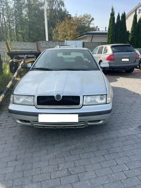 Srebrny samochód osobowy marki Skoda widoczny od przodu, zaparkowany na kostce brukowej. W tle po prawej stronie stoi inny srebrny samochód, a po lewej przyczepa transportowa. W oddali widoczne są drzewa i fragment budynku.