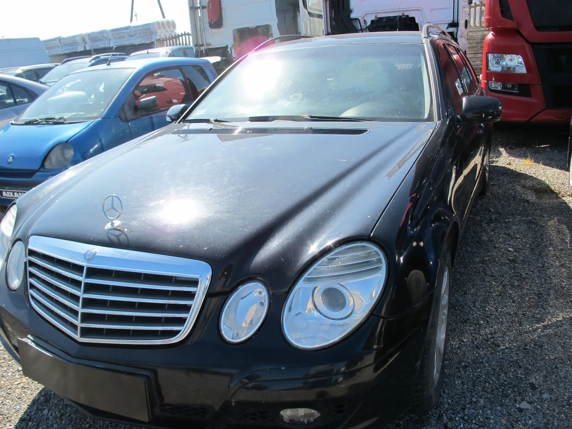 Czarny samochód osobowy marki Mercedes-Benz zaparkowany na żwirowym placu. Widoczny jest przód pojazdu z charakterystycznym grillem i logo na masce. W tle znajdują się inne samochody, w tym niebieski mały samochód oraz czerwony pojazd ciężarowy.