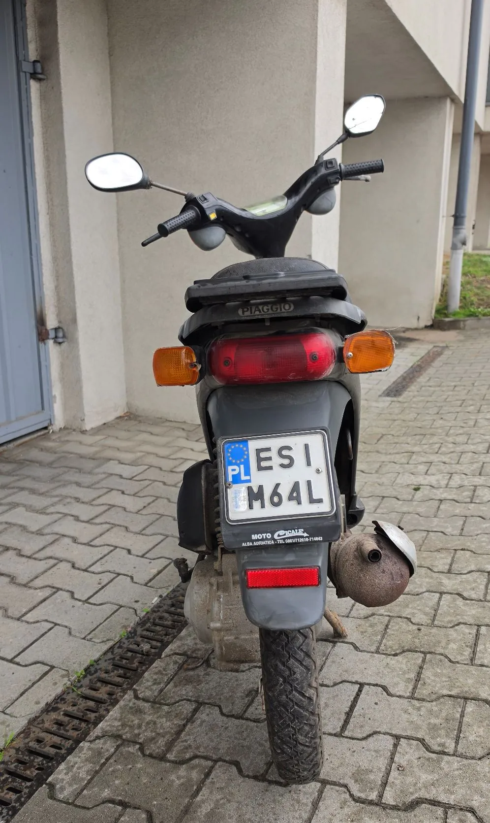 Widok od tyłu na ciemnoszary skuter marki Piaggio zaparkowany na kostce brukowej przed jasną ścianą budynku. Skuter posiada pomarańczowe kierunkowskazy, czerwone światło stopu oraz polską tablicę rejestracyjną o numerze ESI M64L. Widoczne są dwa lusterka wsteczne oraz rura wydechowa po prawej stronie.