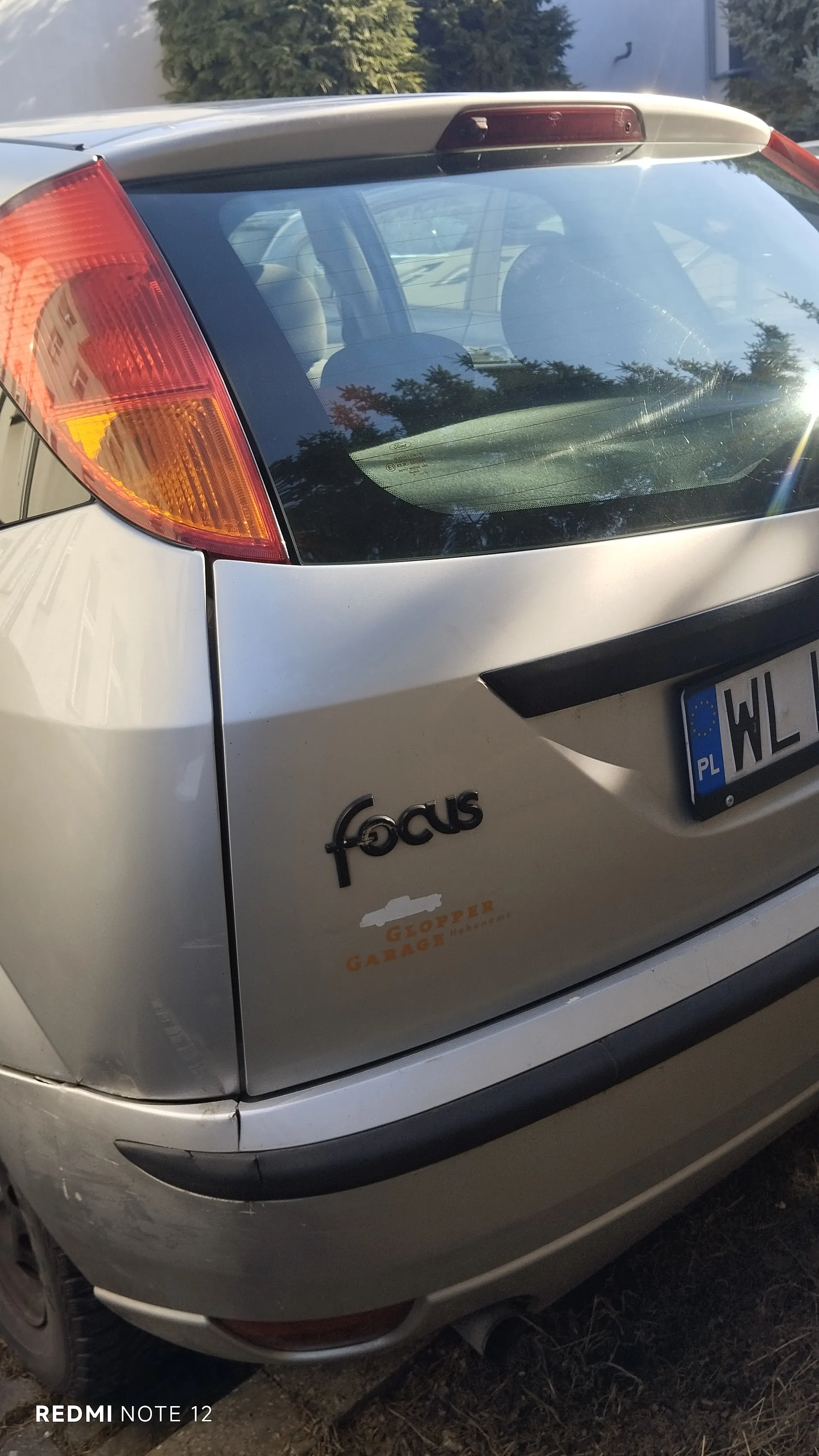 Tył srebrnego samochodu Ford Focus z widocznym napisem "focus" na bagażniku. Na bagażniku naklejka z napisem "Glopper Garage" oraz częściowo widoczna tablica rejestracyjna z oznaczeniem "PL WL". Czerwone i pomarańczowe światła tylnego reflektora, tylne szyby z odbiciem drzew i budynków. Na dole zdjęcia widoczny napis "REDMI NOTE 12". Samochód stoi na podłożu z trawą i ziemią.