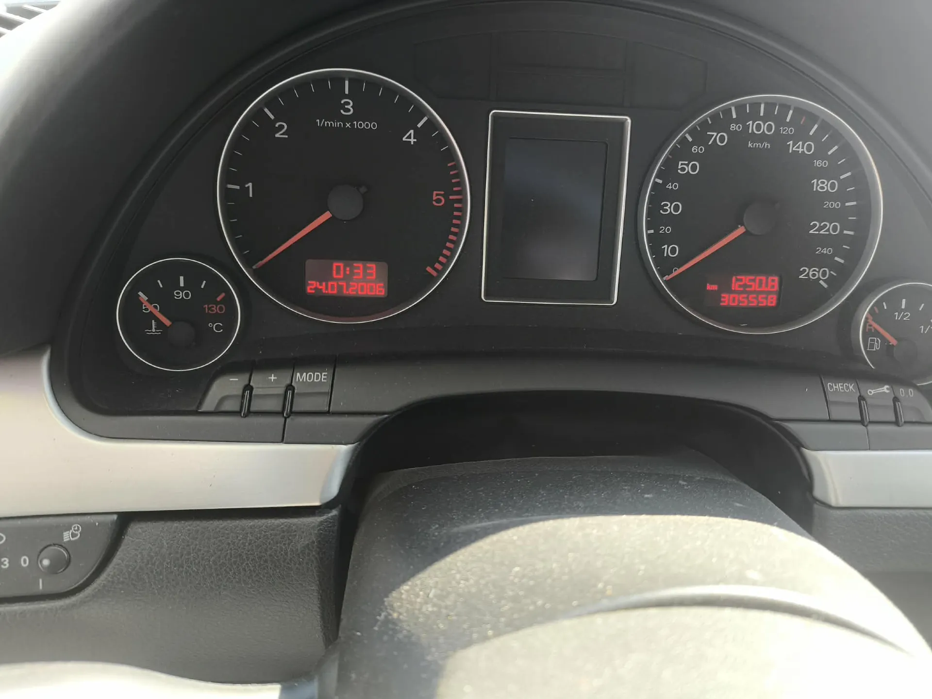 Zbliżenie na deskę rozdzielczą samochodu. Widoczne są trzy analogowe wskaźniki: po lewej temperatura cieczy chłodzącej, w centrum obrotomierz z cyfrowym wyświetlaczem pokazującym godzinę 0:33 i datę 24.07.2006, oraz po prawej prędkościomierz z cyfrowym licznikiem przebiegu wskazującym 305558 km i wartością 1250.8. Po prawej stronie znajduje się również wskaźnik poziomu paliwa. Poniżej zegarów widoczne są przyciski sterujące, w tym przycisk MODE.