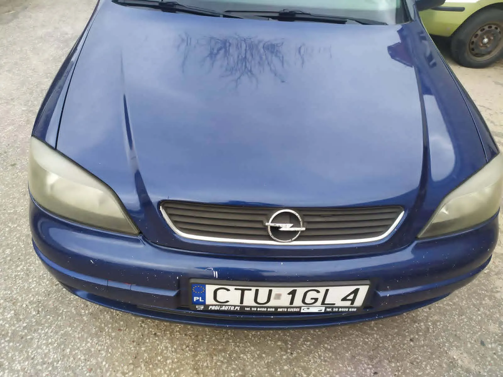 Przód niebieskiego samochodu marki Opel z tablicą rejestracyjną CTU 1GL4. Na tablicy widoczny jest napis PL oraz logo Unii Europejskiej po lewej stronie. Poniżej tablicy znajduje się napis PROFAUTO.PL. Samochód stoi na podłożu z drobnej kostki. W tle po prawej stronie widoczna jest część żółtego pojazdu z kołem.