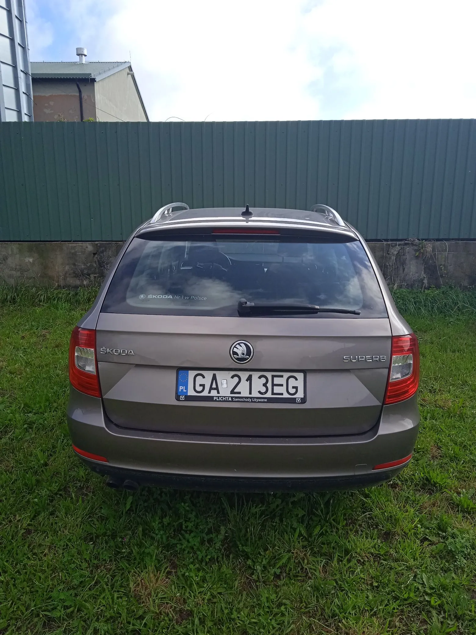 Tylny widok brązowego samochodu marki Skoda Superb zaparkowanego na trawie przed zielonym, falistym płotem. Samochód ma polską tablicę rejestracyjną o numerze GA 213EG. Na ramce tablicy widnieje napis 'PLICHTA Samochody Używane'. Na tylnej szybie znajduje się naklejka z tekstem 'SKODA Nr 1 w Polsce'.