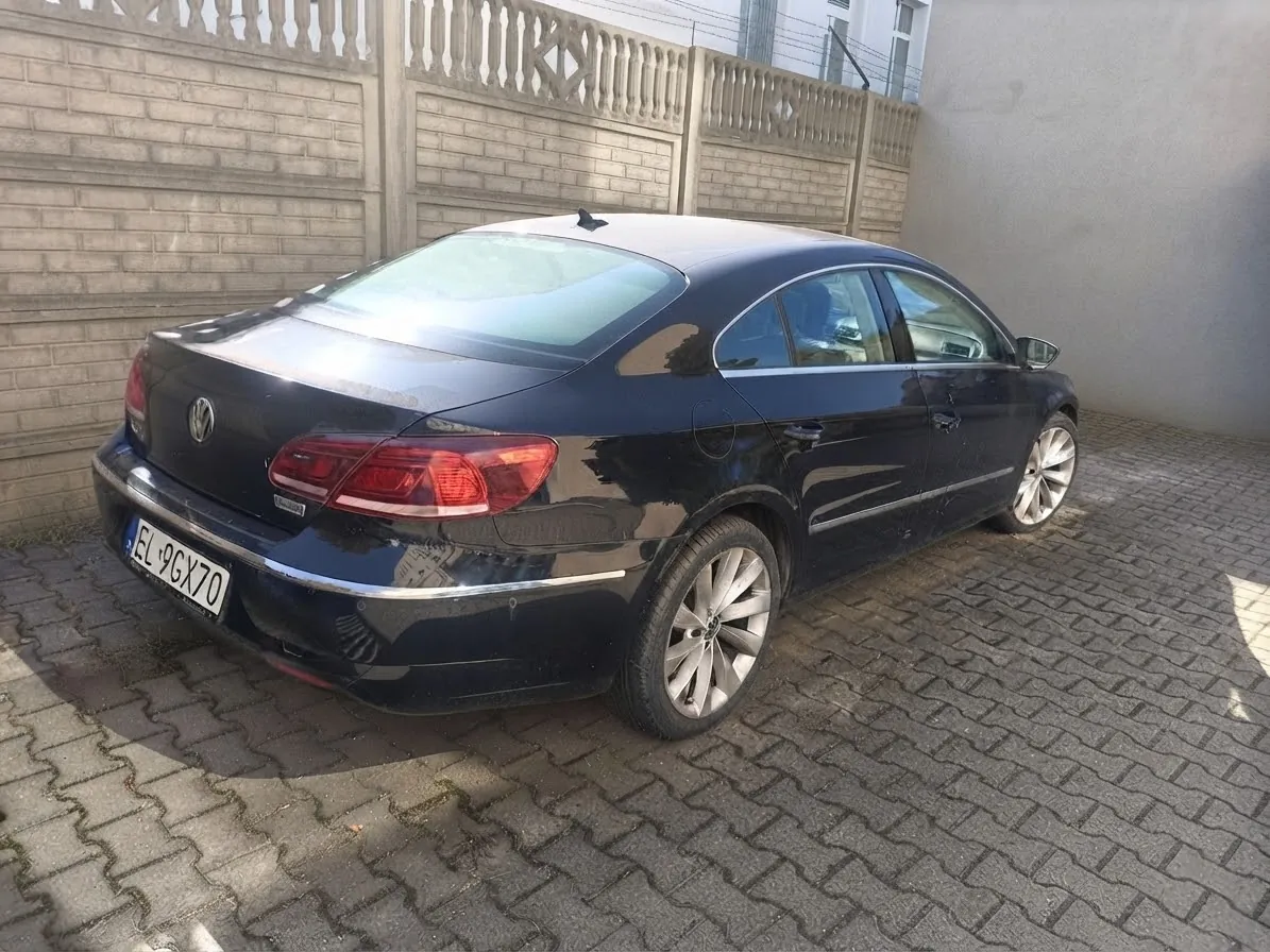 Czarny samochód osobowy marki Volkswagen zaparkowany tyłem na kostce brukowej. Pojazd posiada tablicę rejestracyjną o numerze EL 9GX70. W tle widoczny jest szary betonowy płot oraz fragment jasnej ściany budynku.