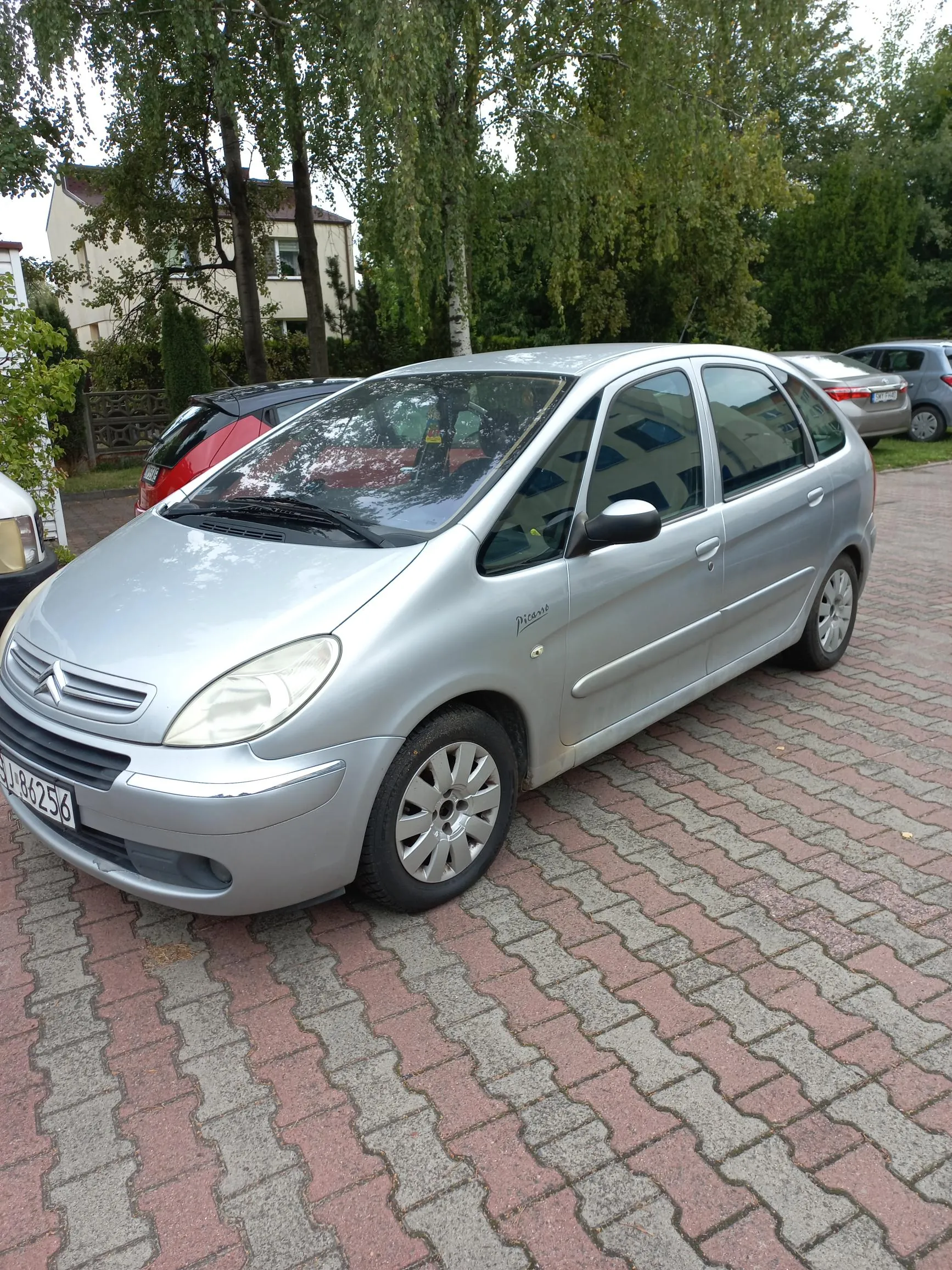 Srebrny samochód marki Citroën Picasso zaparkowany na kostce brukowej. W tle widoczne są inne samochody, zielone drzewa oraz jasny budynek mieszkalny. Na masce samochodu widoczne jest logo Citroëna, a na boku napis 'Picasso'. Tablica rejestracyjna zaczyna się od liter 'BJ'.
