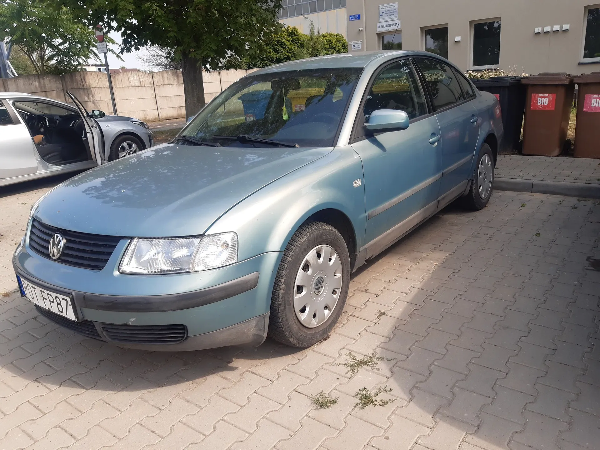 Niebieski samochód osobowy marki Volkswagen zaparkowany na kostce brukowej. Pojazd jest ustawiony pod kątem, widoczny jest przód i prawy bok auta. Na przednim zderzaku znajduje się tablica rejestracyjna o numerze FP 87. W tle widoczny jest inny, srebrny samochód z otwartymi drzwiami, zielone drzewa oraz fragment jasnego budynku z oknami i brązowymi pojemnikami na odpady z napisem BIO.
